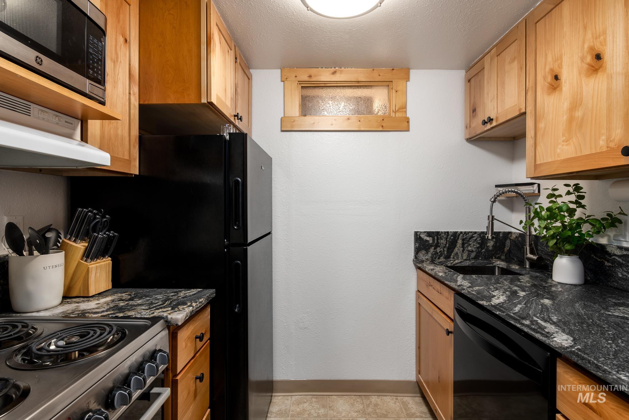 Compact kitchen featuring light wood cabinetry, black appliances, a stainless steel microwave, a dual-basin sink with a gooseneck faucet, and dark granite countertops