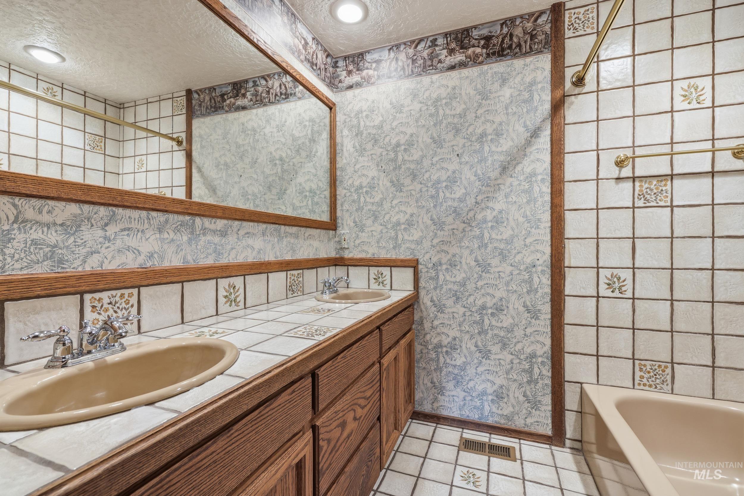 Bathroom with a textured ceiling, wallpapered walls, double vanity, light tile patterned flooring, and  shower combination