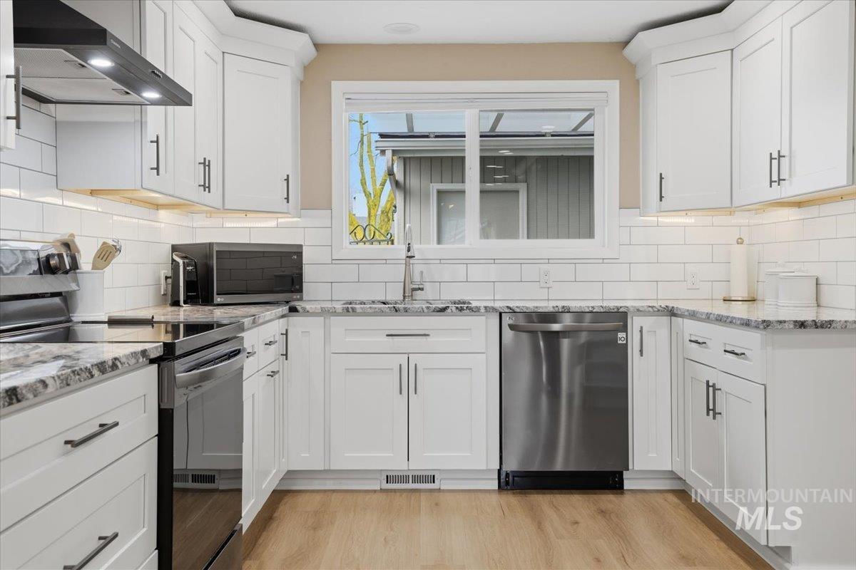 Kitchen with appliances with stainless steel finishes, wall chimney range hood, light stone countertops, and white cabinets
