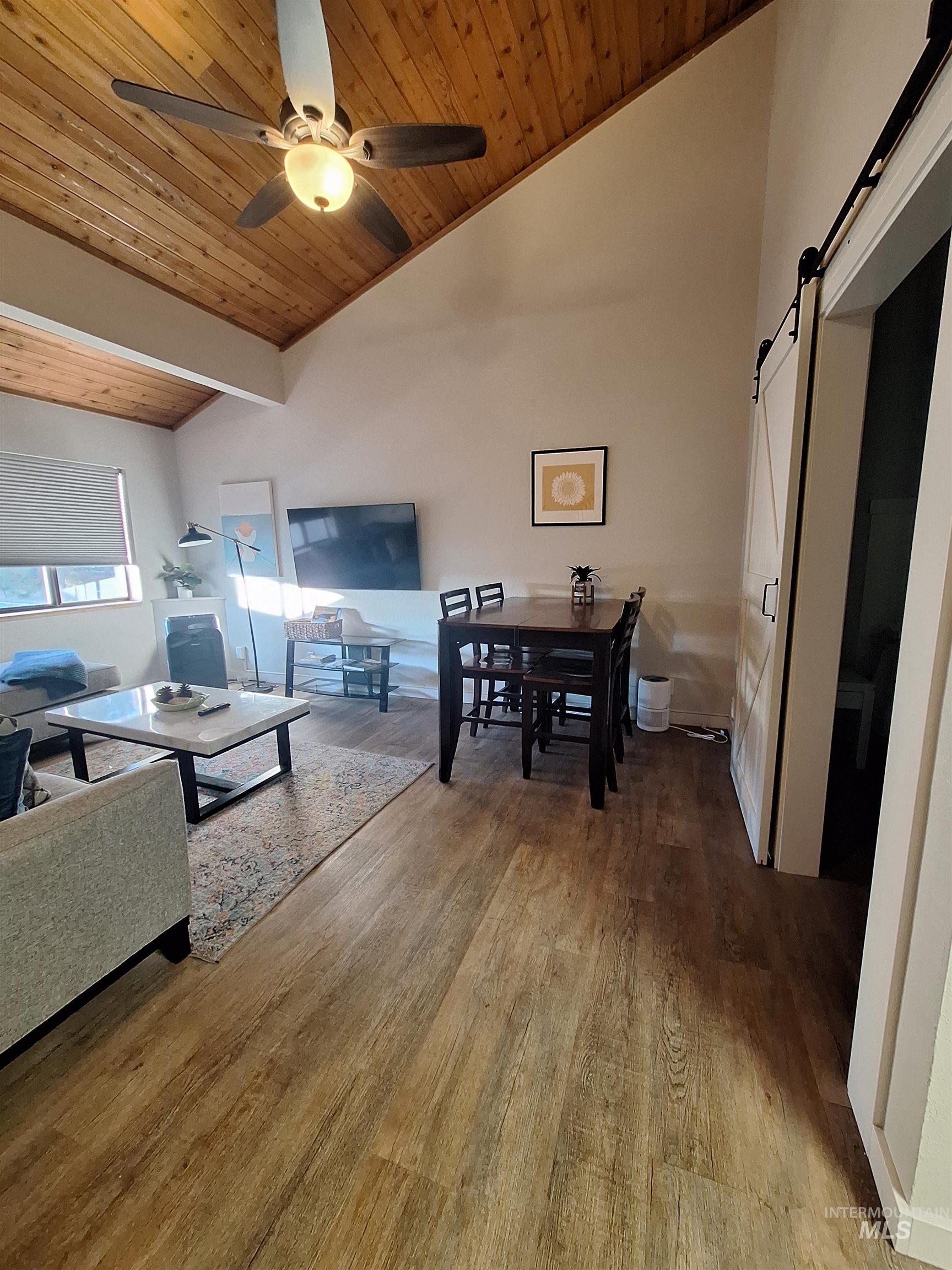 Living room with wood ceiling, vaulted ceiling, light wood finished floors, a barn door, and a ceiling fan