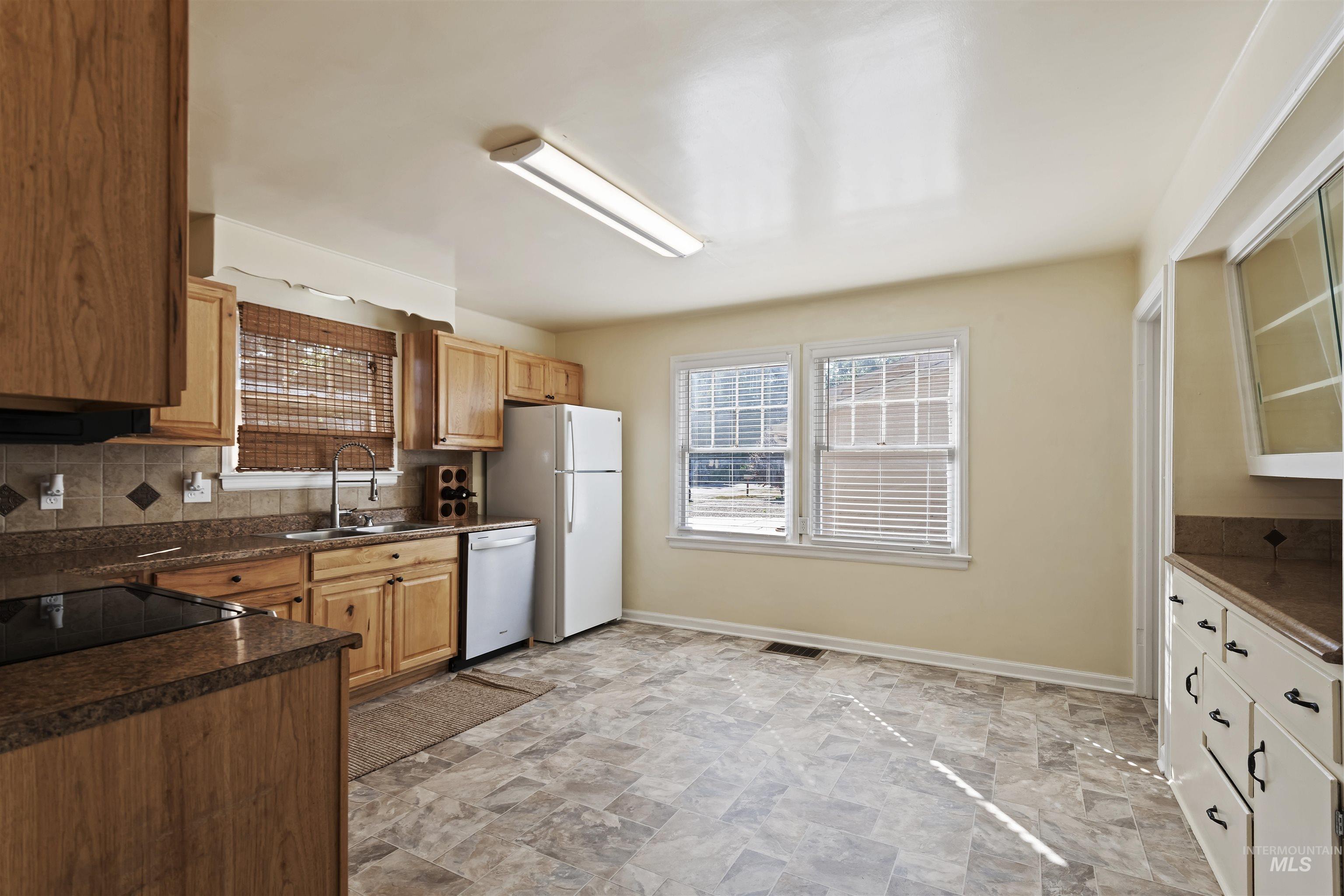 Kitchen with dark countertops, backsplash, stone finish flooring, white dishwasher, and glass insert cabinets