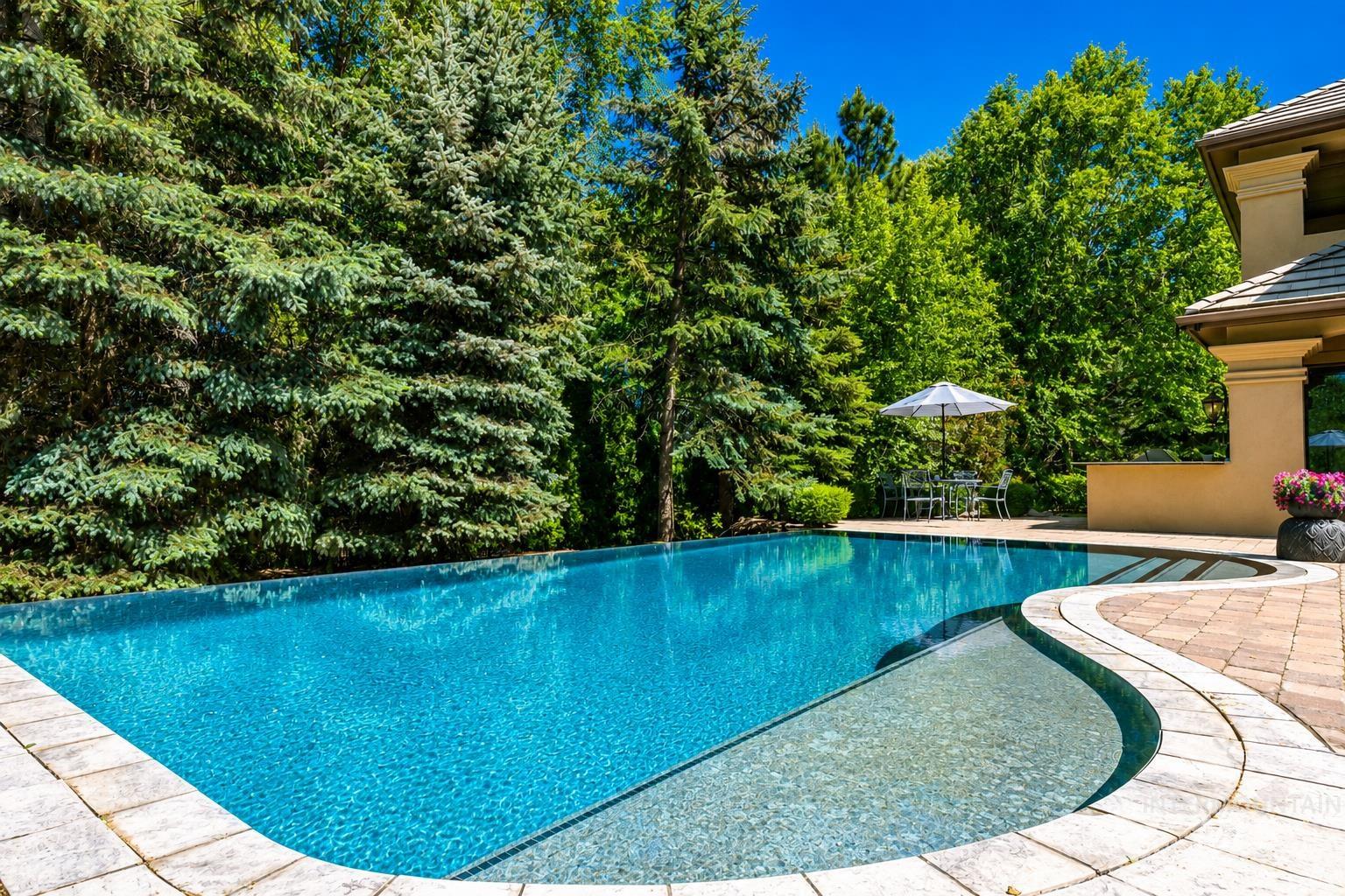 Curved swimming pool with an integrated shallow entry area, surrounded by light-toned paver decking and mature evergreen landscaping