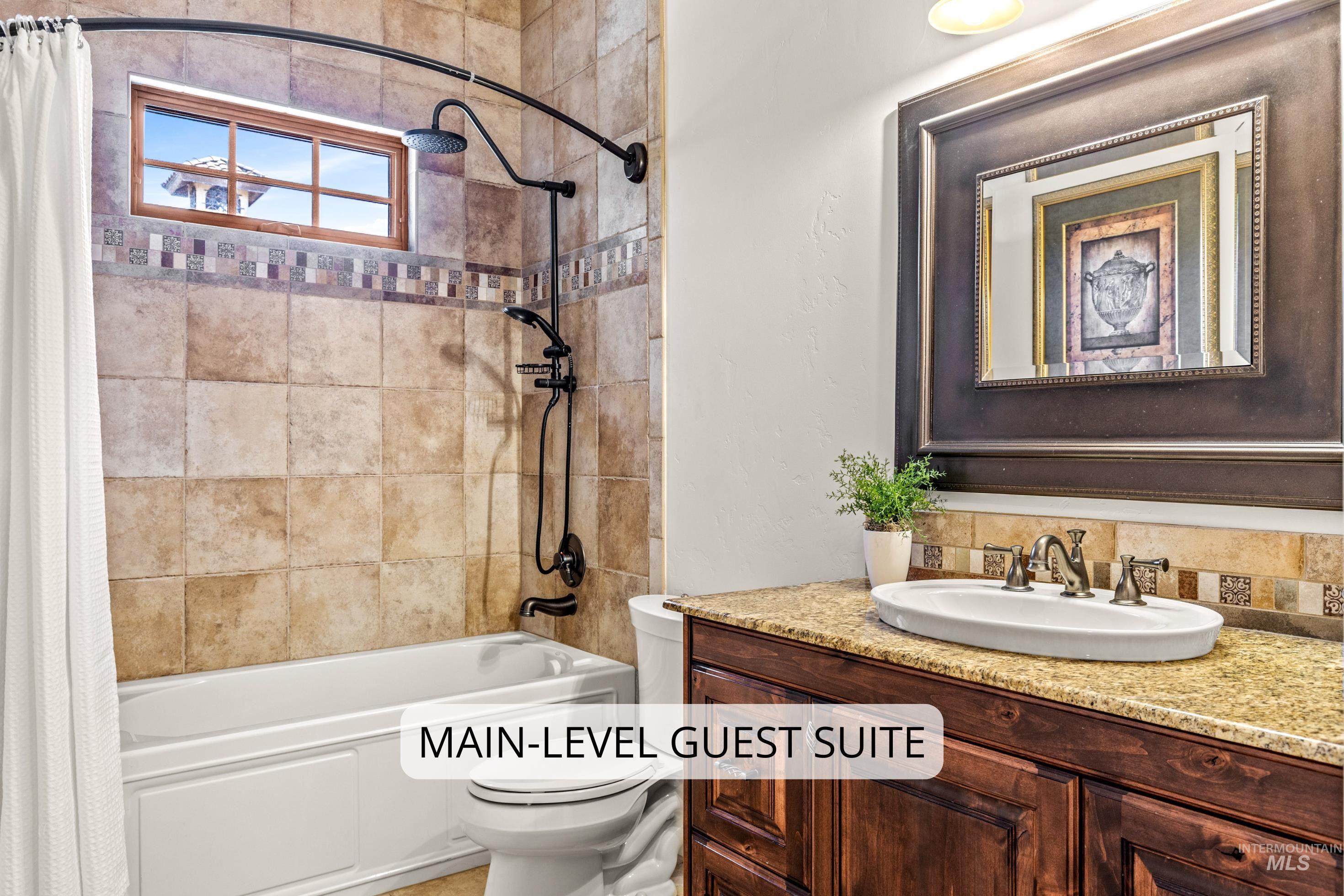 Bathroom featuring a full-size bathtub with an overhead shower and handheld showerhead, natural stone-look wall tiling with a decorative accent band, a vanity with a granite-style countertop, a rectangular vessel sink, and a framed mirror