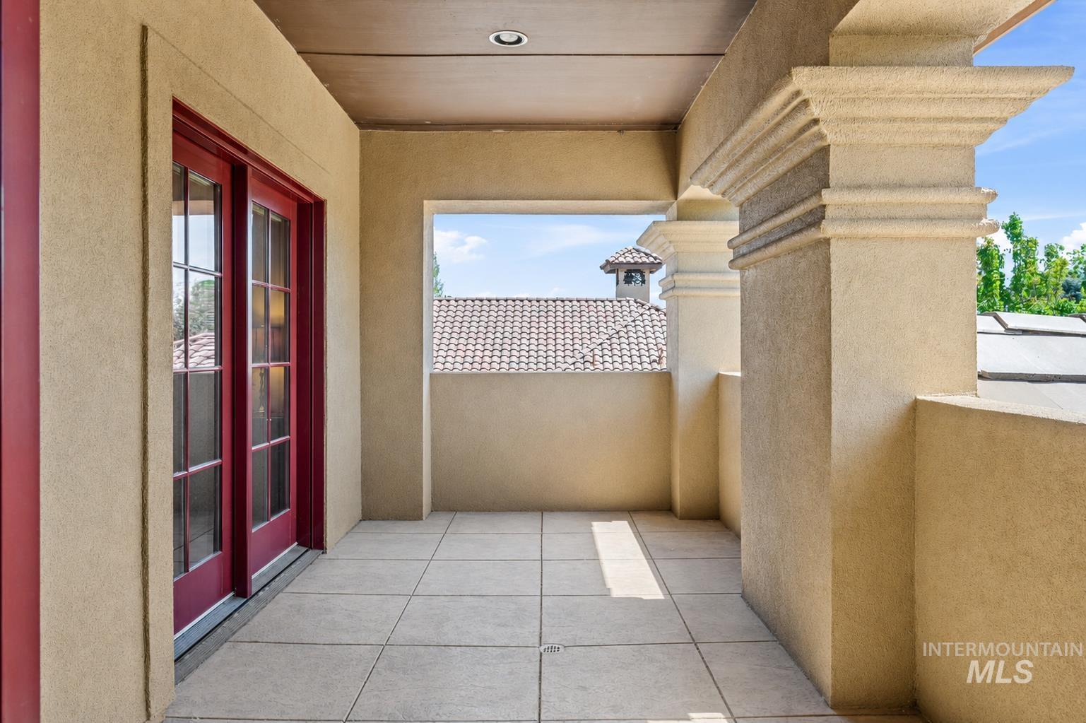 Balcony featuring light-toned tile flooring, stucco walls, decorative architectural columns, and red framed glass-paneled double doors