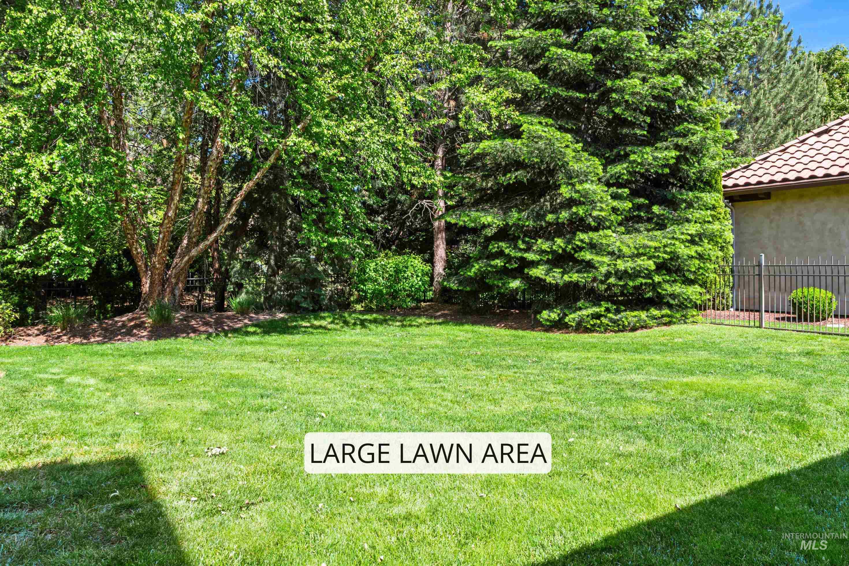 Expansive green lawn area featuring mature trees, diverse plantings, and a partial view of a stucco exterior with a terracotta tile roof and black metal fencing