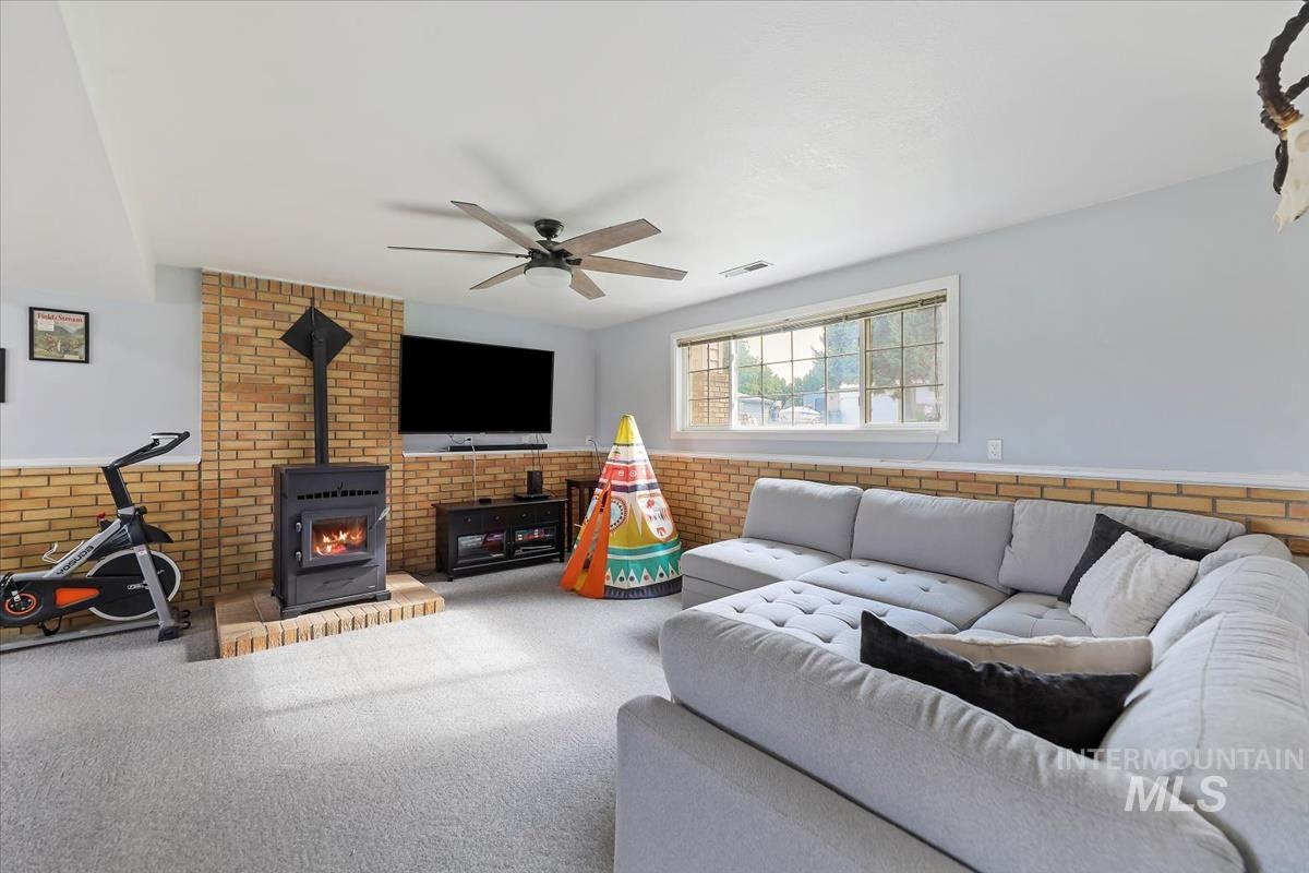 Living room with a wood stove, wainscoting, ceiling fan, carpet flooring, and brick wall