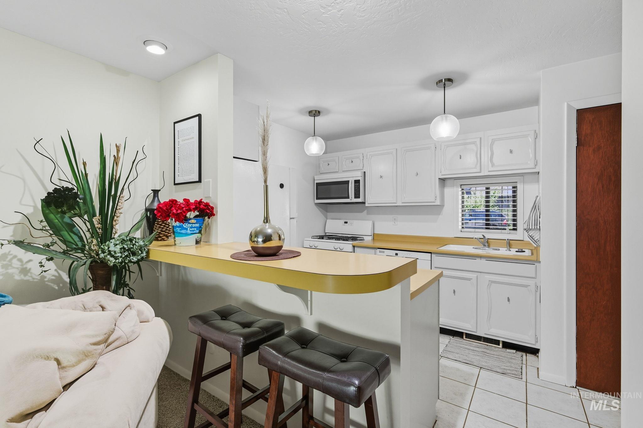 Kitchen featuring light countertops, white cabinetry, white appliances, hanging light fixtures, and a kitchen bar