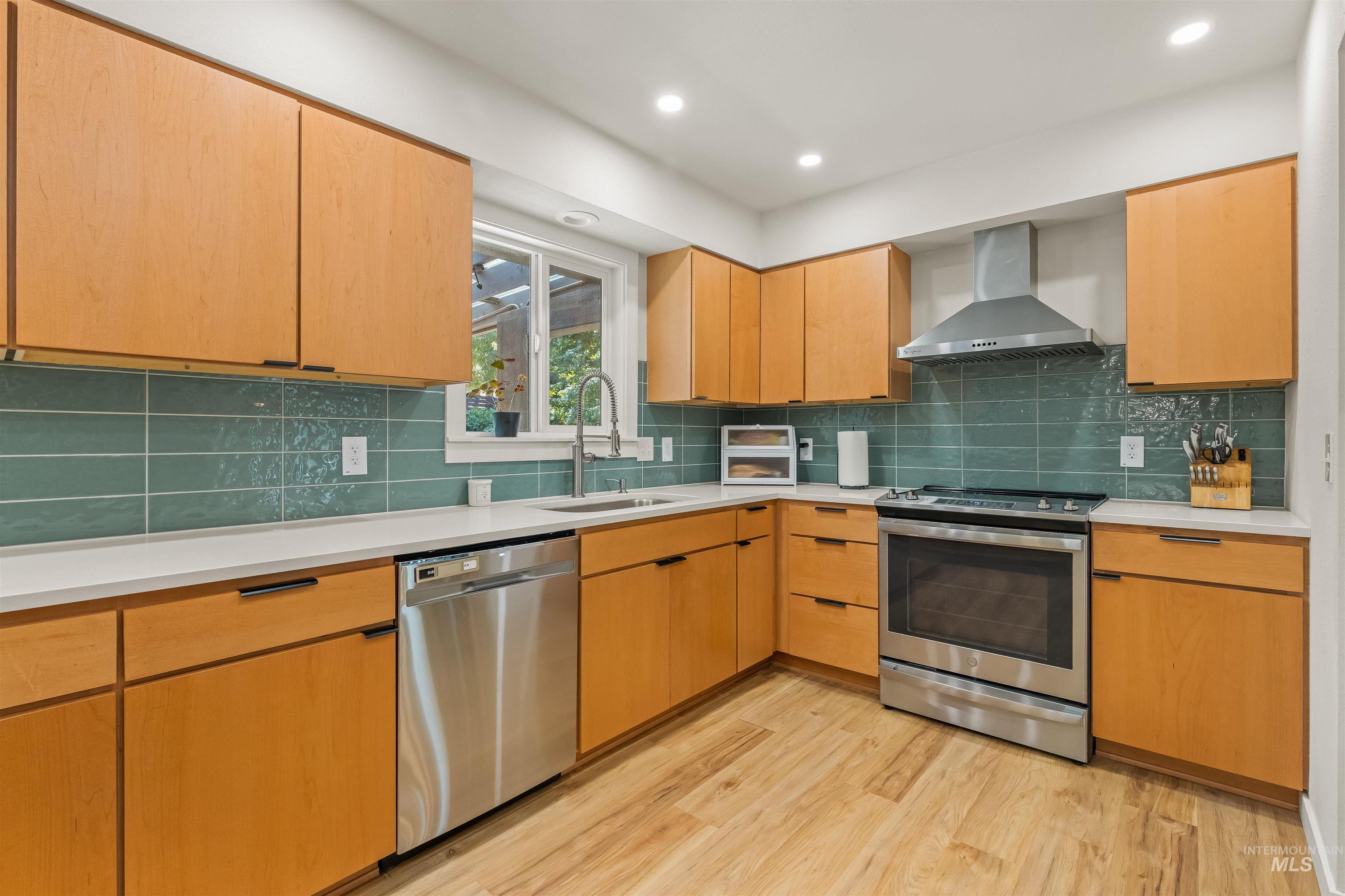 Kitchen featuring wood-finish cabinetry, stainless steel appliances, a green tile backsplash, light-toned countertops, and wood-finish flooring