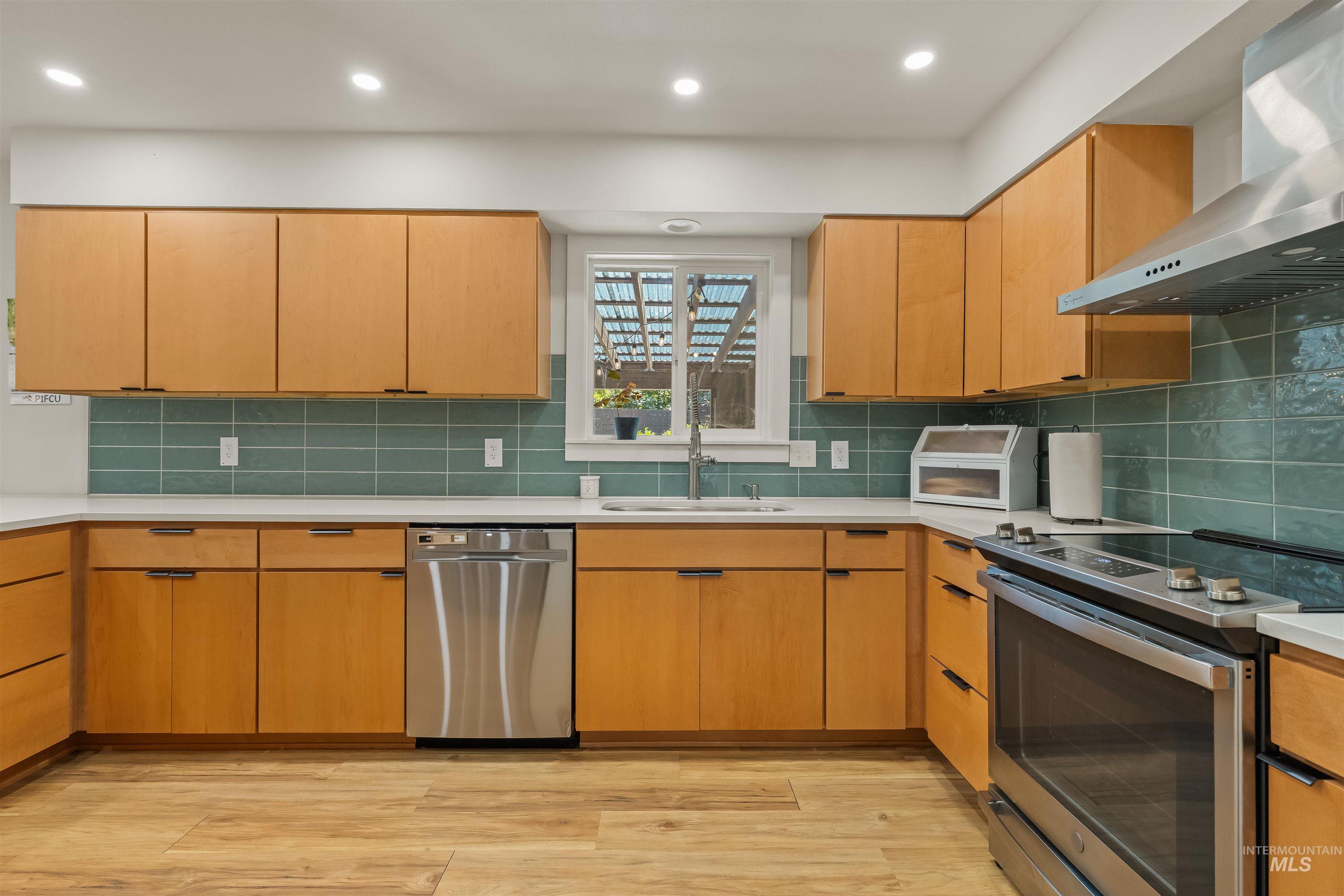 Kitchen featuring light wood cabinetry, white countertops, stainless steel appliances, a teal subway tile backsplash, and wood-finish flooring