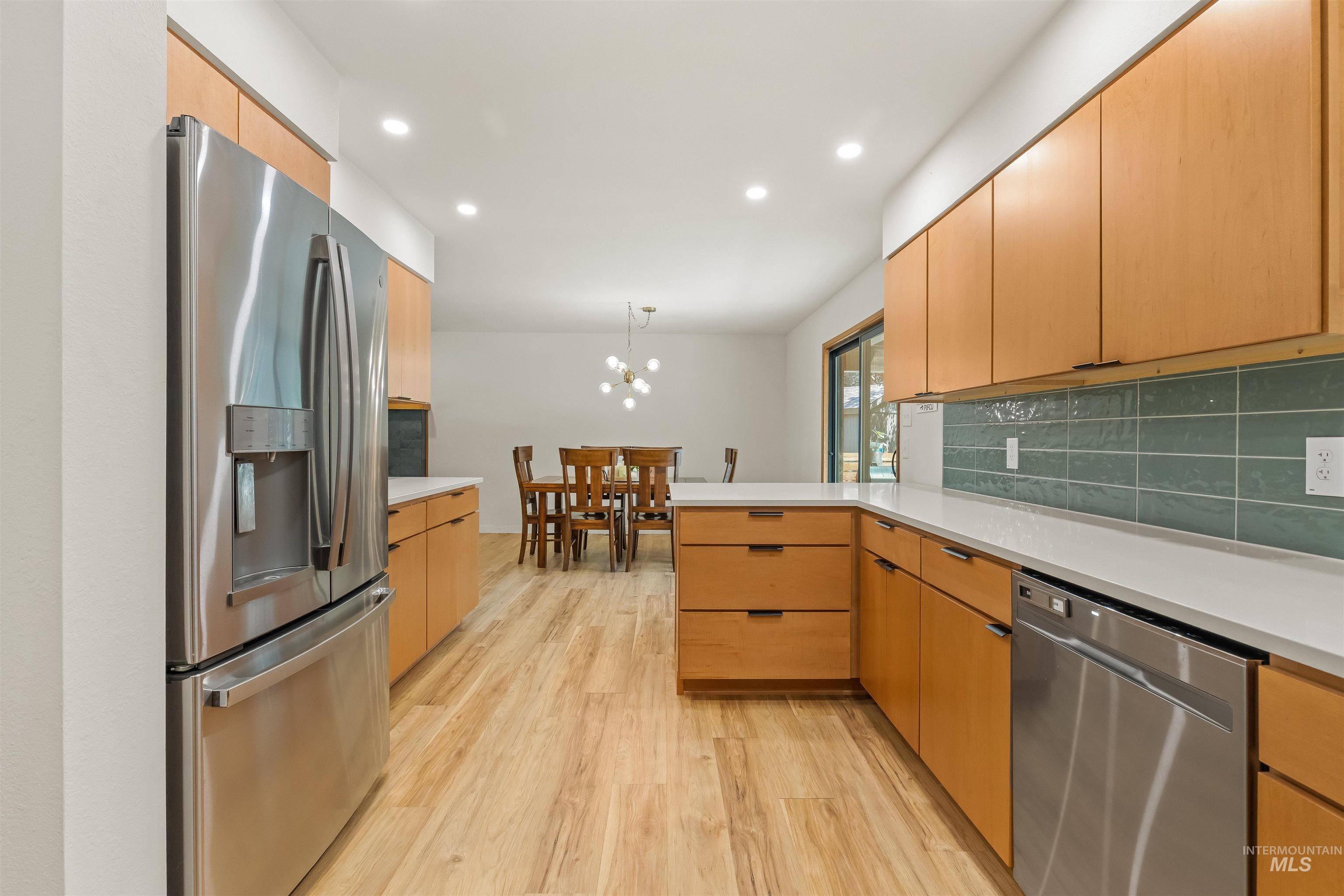 Kitchen with wood-finish flooring, stainless steel appliances, light wood cabinetry, white countertops, and a teal tile backsplash