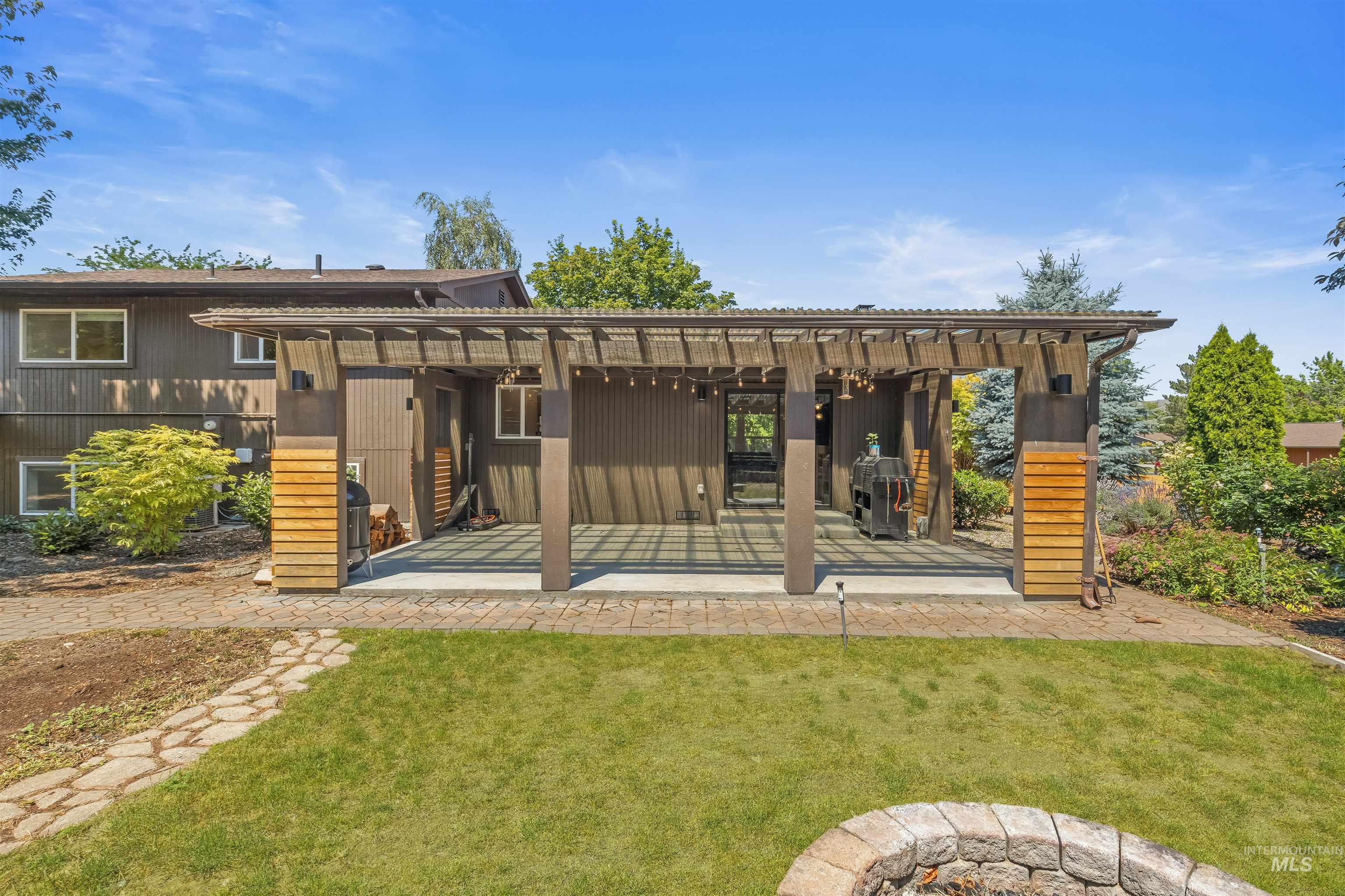 Expansive backyard featuring a covered patio with string lighting, a stone paver patio, lush green lawn, and a stone fire pit