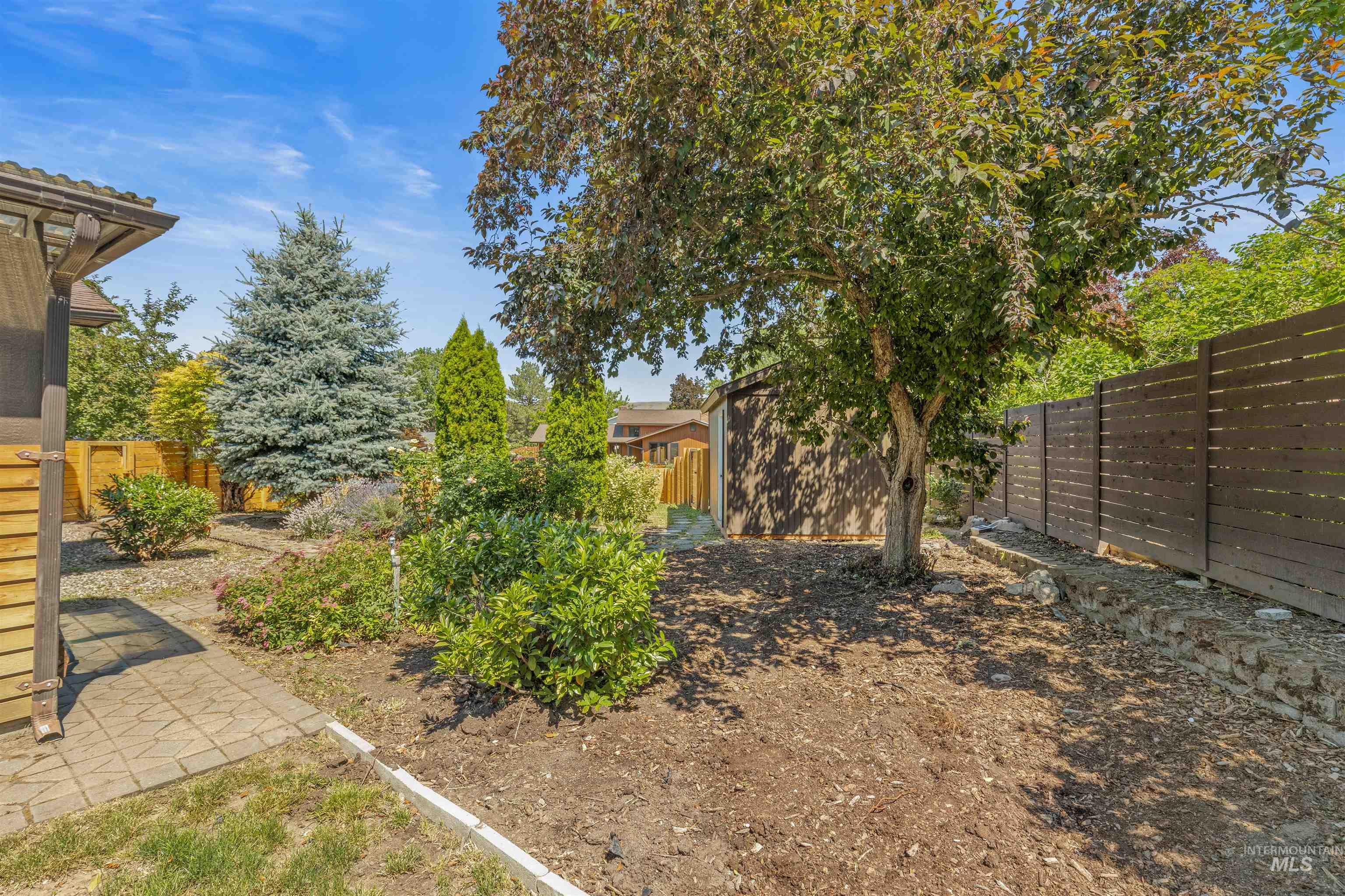 Landscaped backyard featuring established trees and shrubs, wood fencing, and a paved patio area
