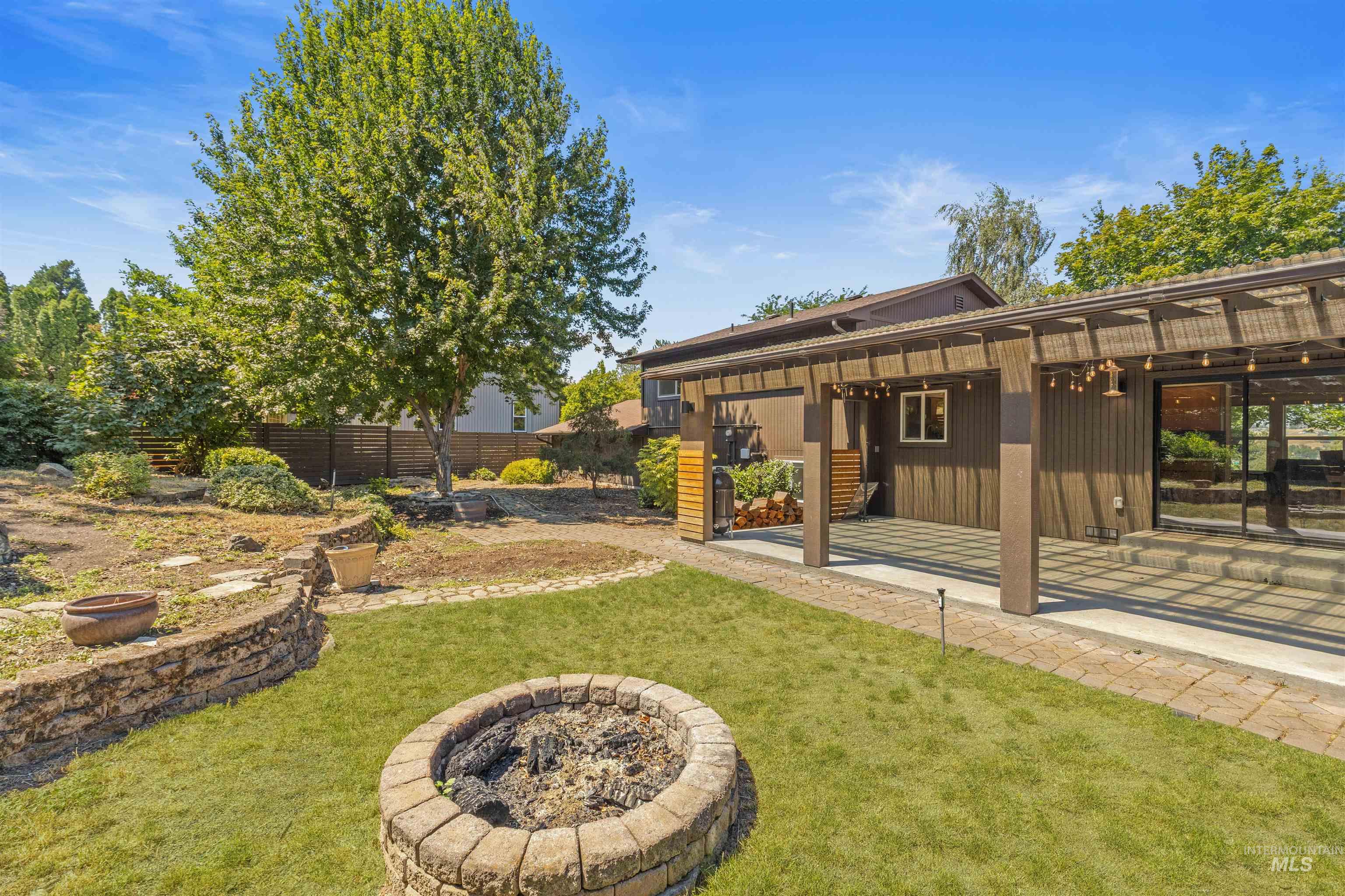 Spacious backyard featuring a built-in stone fire pit, tiered landscaping with stone retaining walls, and a large covered patio with string lighting