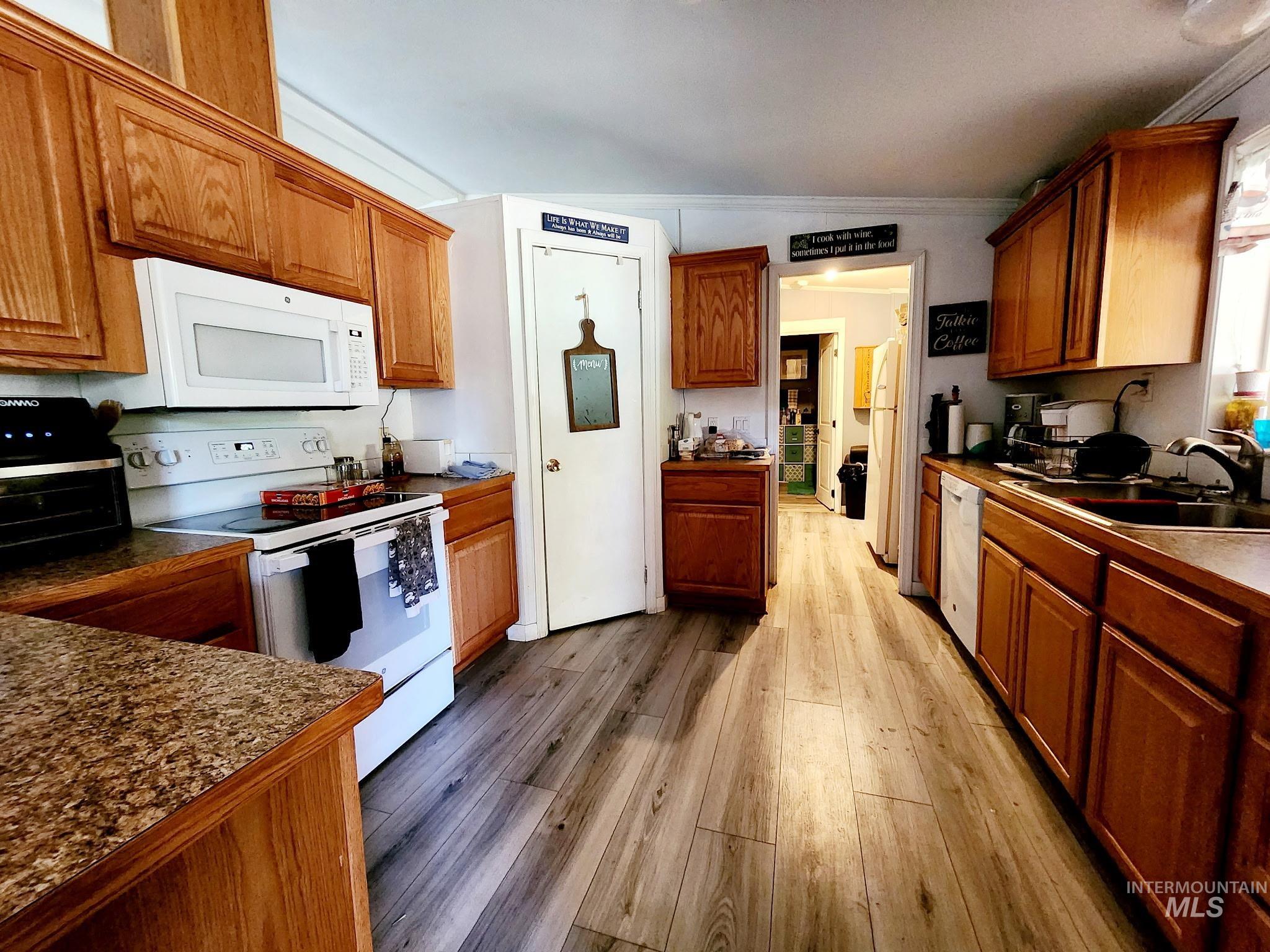 Kitchen with white appliances, ornamental molding, light wood-type flooring, brown cabinetry, and dark countertops