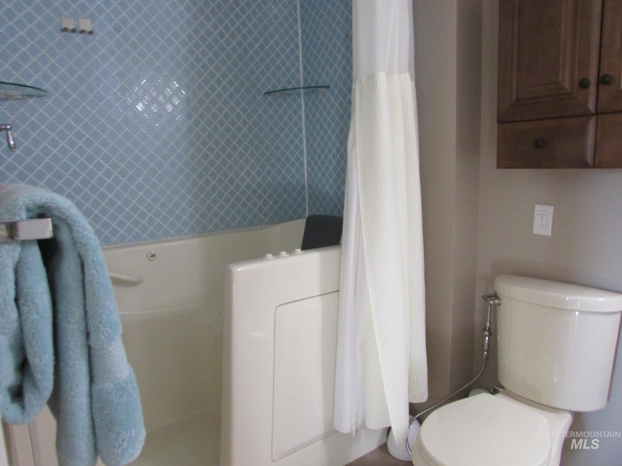 Bathroom featuring a walk-in tub with grab bar, blue geometric wall tiling, a wall-mounted glass shelf, and a white toilet