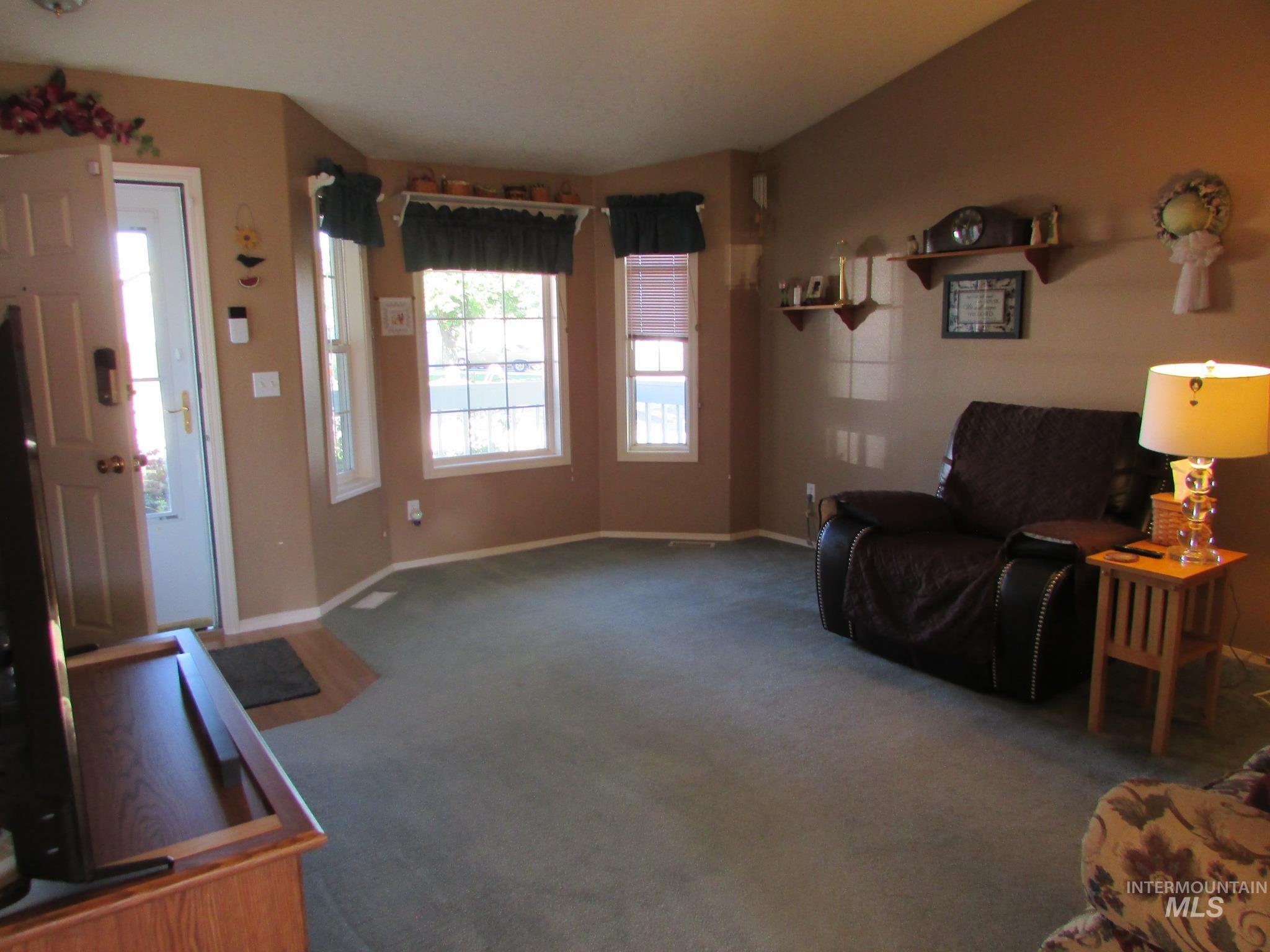Living area featuring bay windows with blinds and valances, light tan wall paint, and green carpet flooring