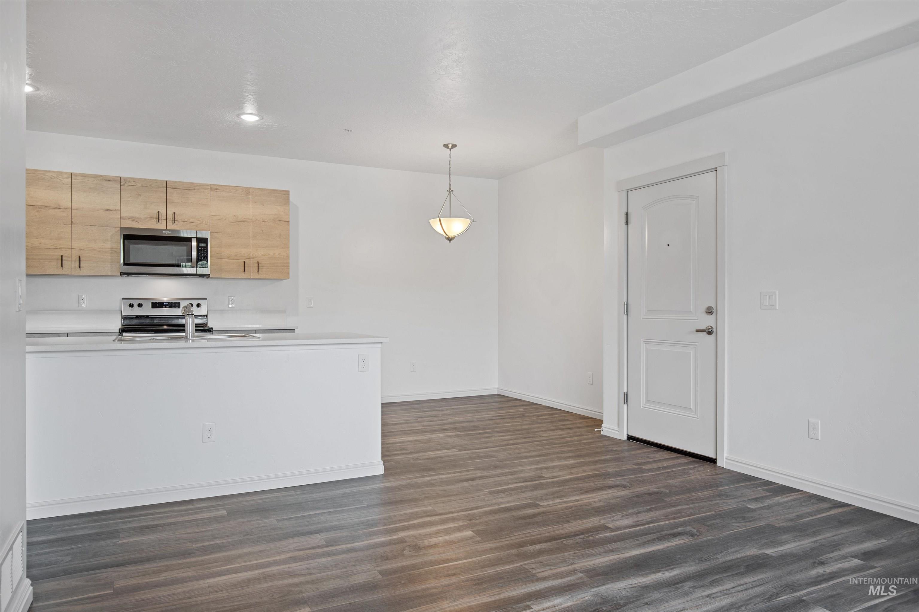 Kitchen featuring stainless steel appliances, dark wood-type flooring, light countertops, decorative light fixtures, and light brown cabinetry
