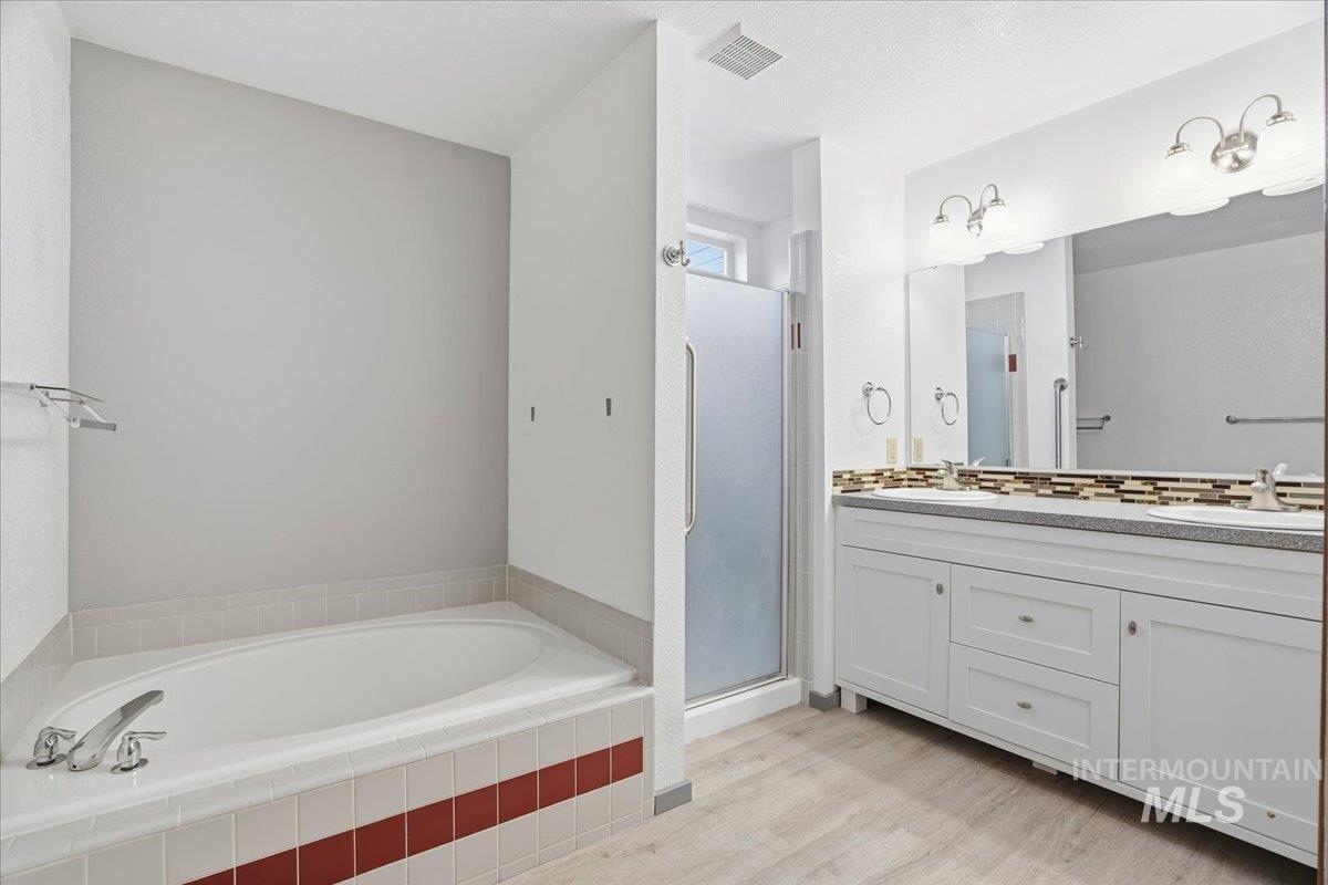 Full bath featuring double vanity, a shower stall, a garden tub, decorative backsplash, and light wood-type flooring