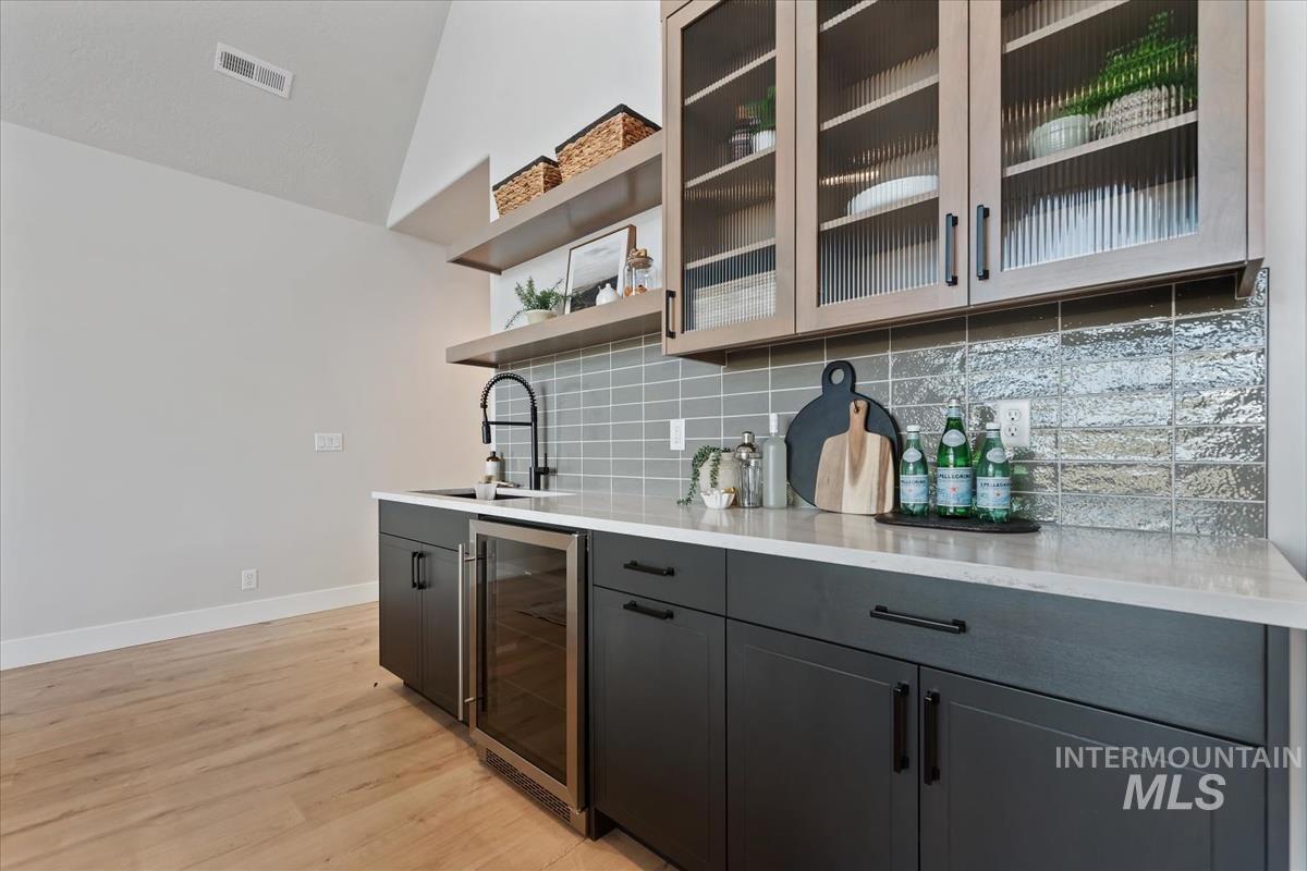 Indoor bar with tasteful backsplash, beverage cooler, open shelves, light wood finished floors, and light stone countertops