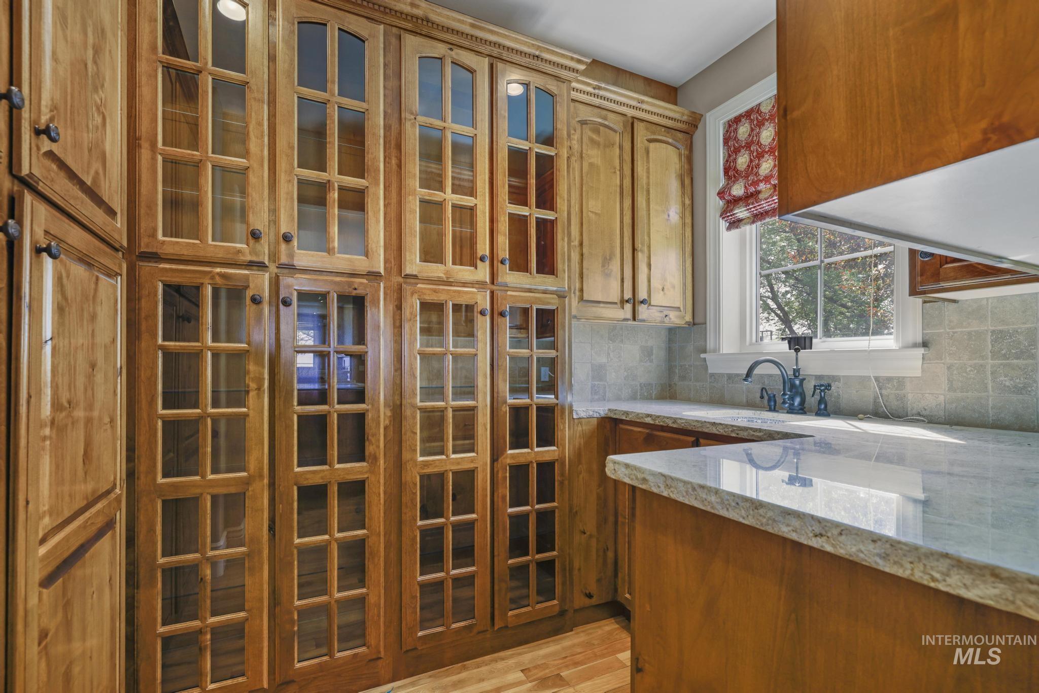 Kitchen featuring light wood-style floors, light stone countertops, decorative backsplash, glass insert cabinets, and brown cabinetry
