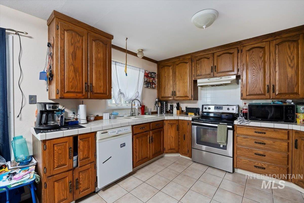 Kitchen with brown cabinetry, stainless steel range with electric cooktop, tile counters, dishwasher, and light tile patterned floors