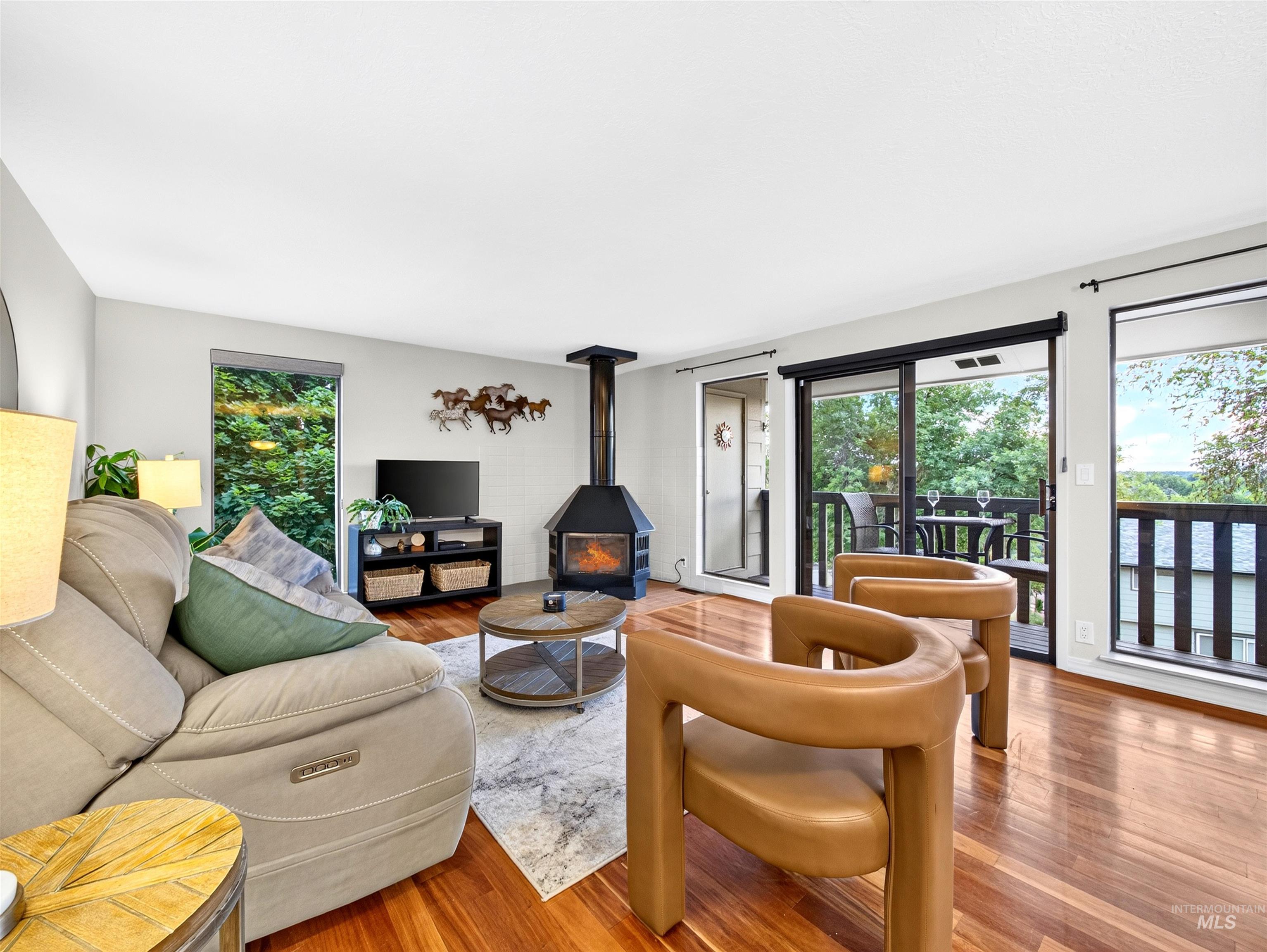 Living area featuring wood-finish flooring, a freestanding fireplace, a sliding glass door to an exterior balcony, a large window offering outdoor scenery, and light-toned walls