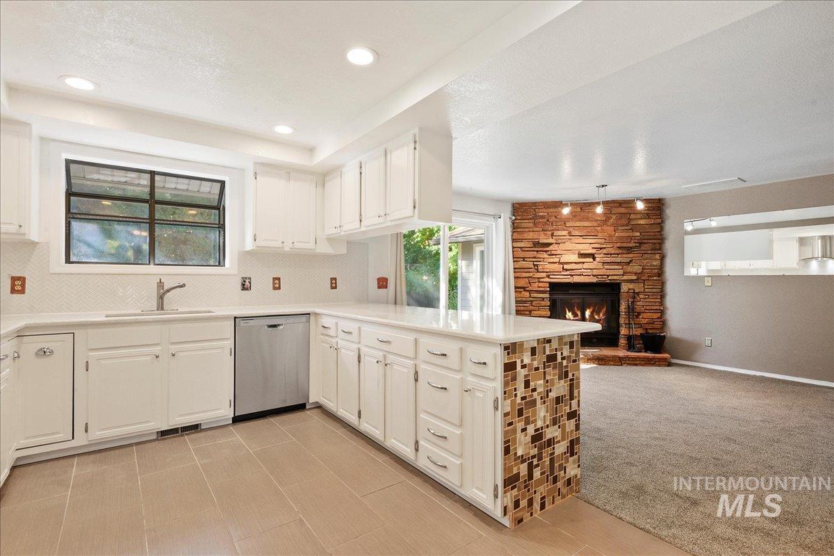 Kitchen with a stone fireplace, stainless steel dishwasher, a peninsula, light carpet, and a textured ceiling
