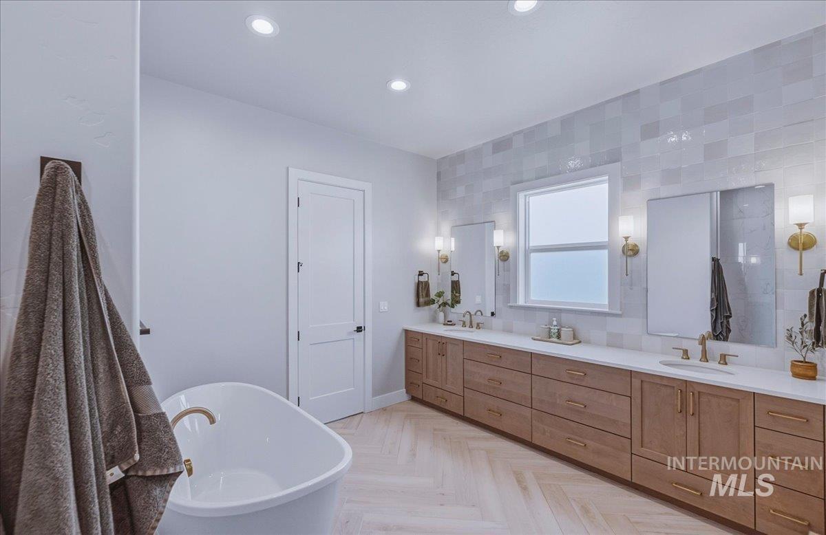 Spacious bath featuring a freestanding soaking tub with brass fixtures, a double vanity with wood-finish cabinetry and white countertops, a tiled accent wall, and herringbone wood-finish flooring