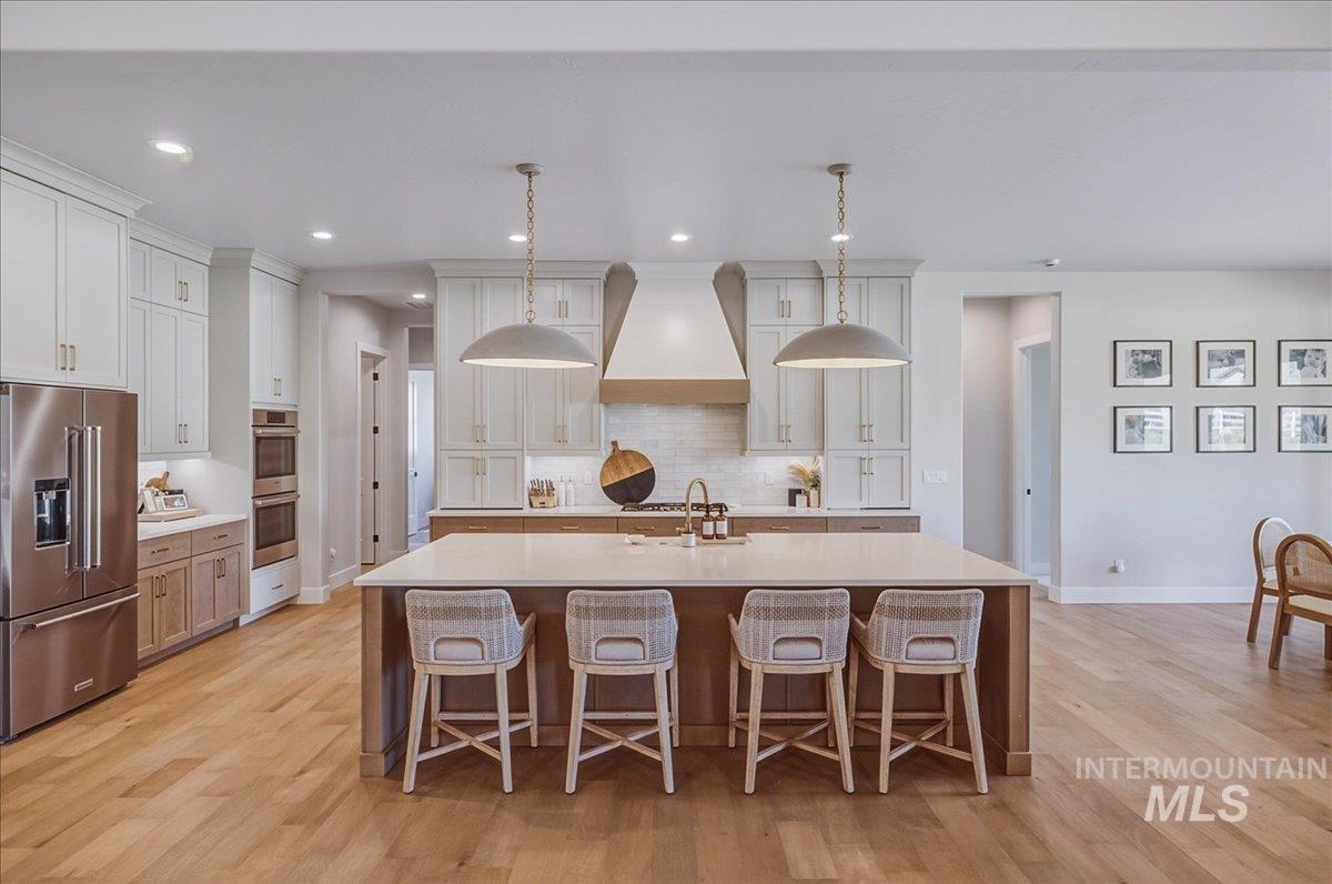 Open-concept kitchen featuring an oversized island with seating, stainless steel appliances, light-toned cabinetry, wood-finish flooring, and pendant lighting