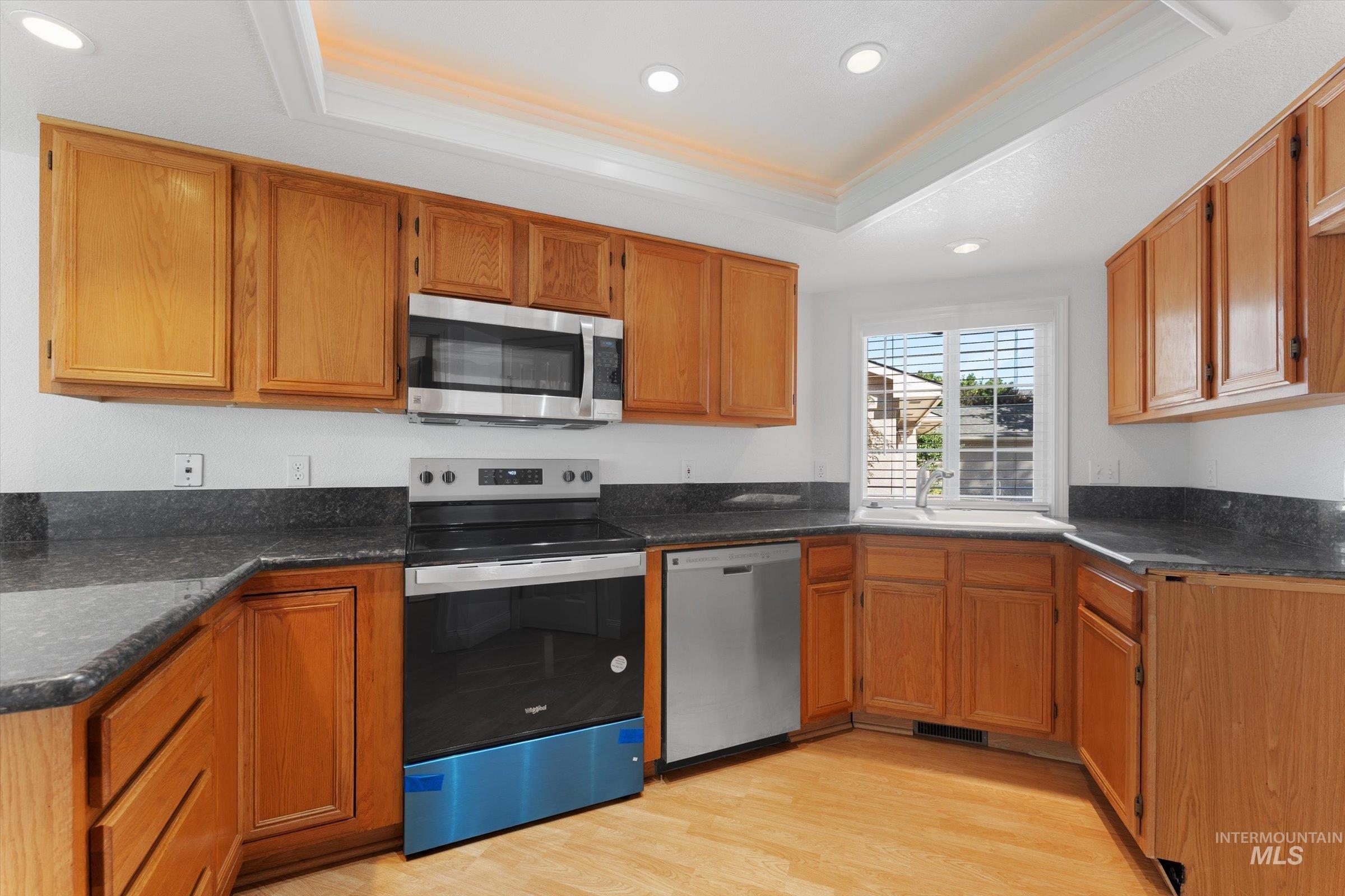Kitchen with a tray ceiling, appliances with stainless steel finishes, light wood finished floors, brown cabinetry, and recessed lighting