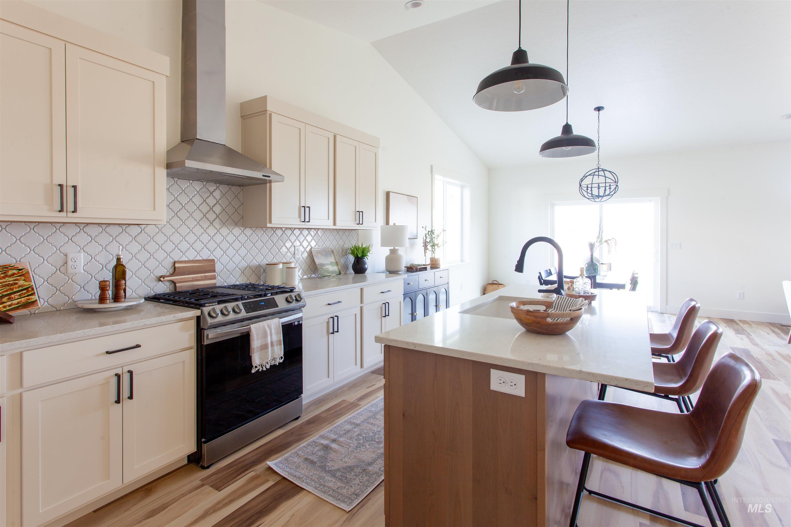 Kitchen with wall chimney range hood, stainless steel range with gas cooktop, a center island with sink, light wood-type flooring, and decorative backsplash