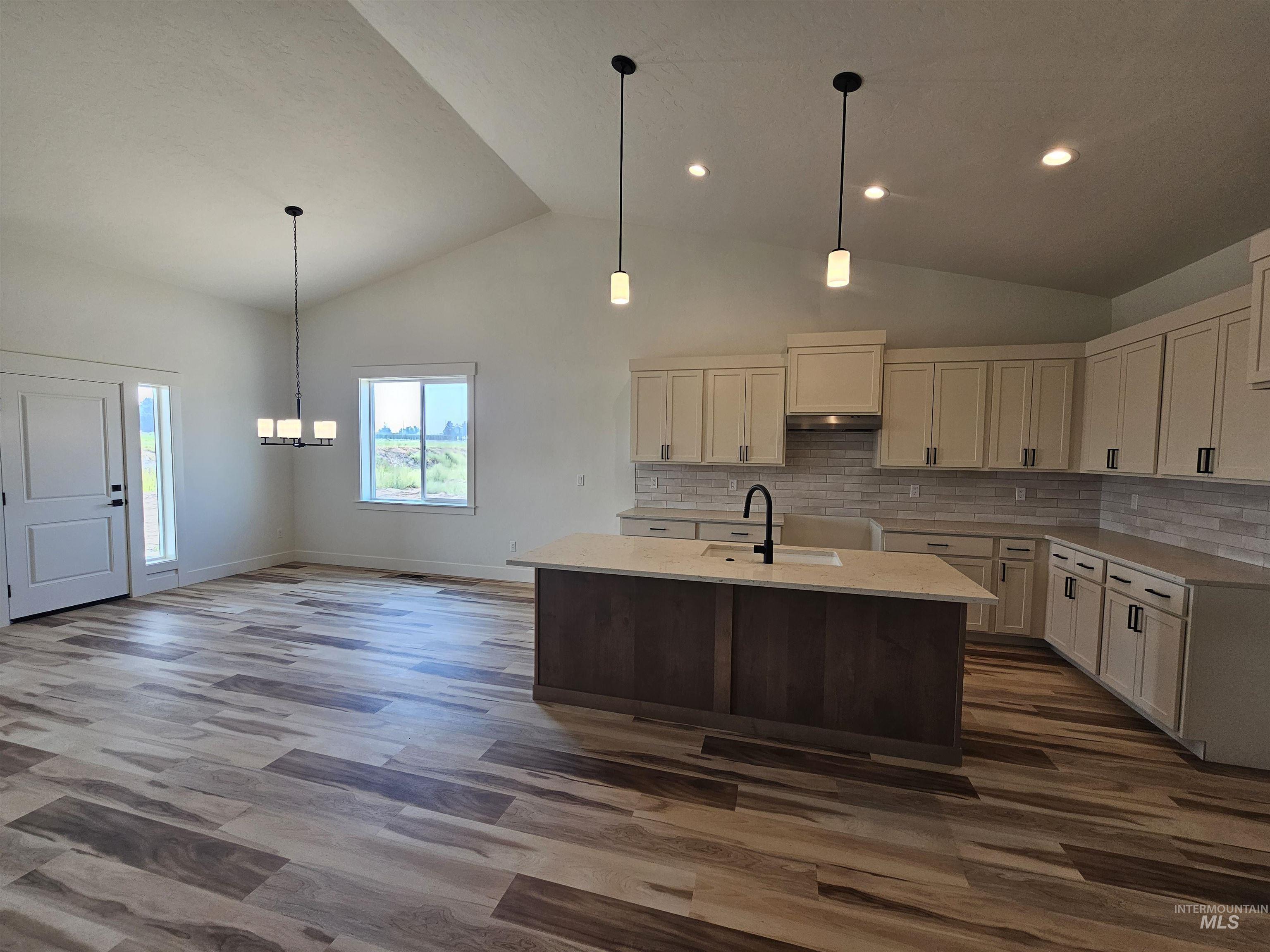 Kitchen with backsplash, a center island with sink, hanging light fixtures, light stone counters, and dark wood finished floors