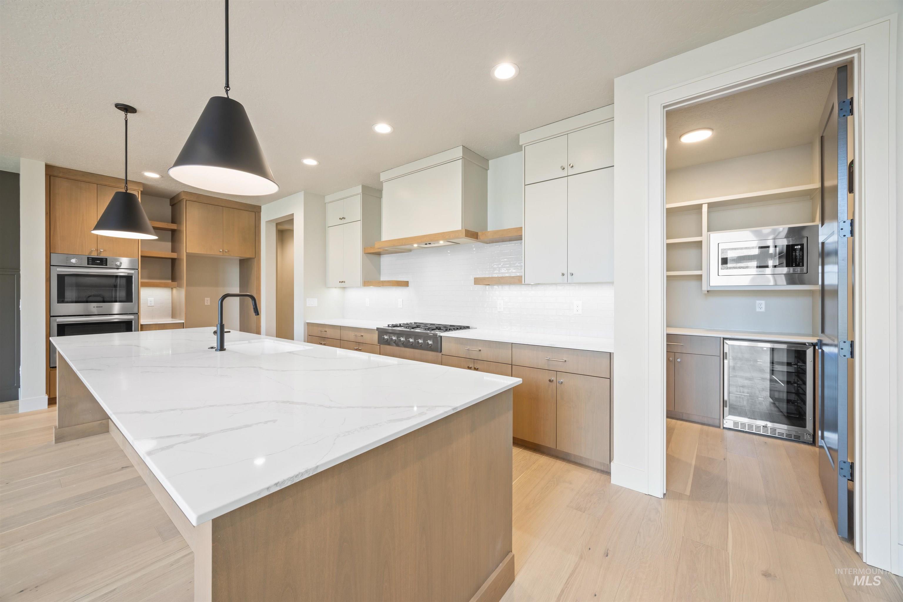 Two tone kitchen featuring open shelves, wine cooler, pendant lighting, light wood-style flooring, and a kitchen island with sink