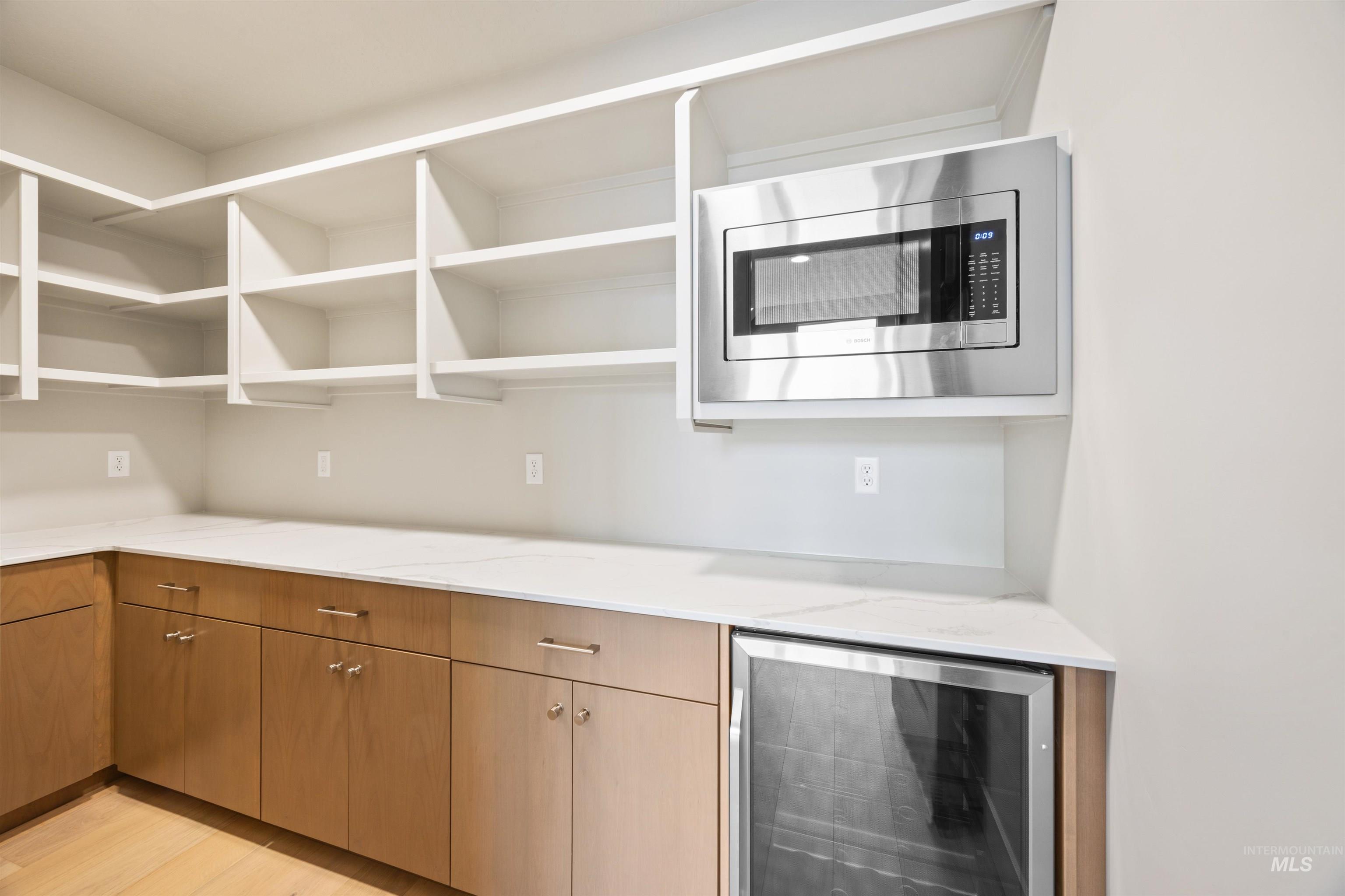 Kitchen with beverage cooler, stainless steel microwave, light wood-type flooring, wood finish cabinetry, and light stone countertops