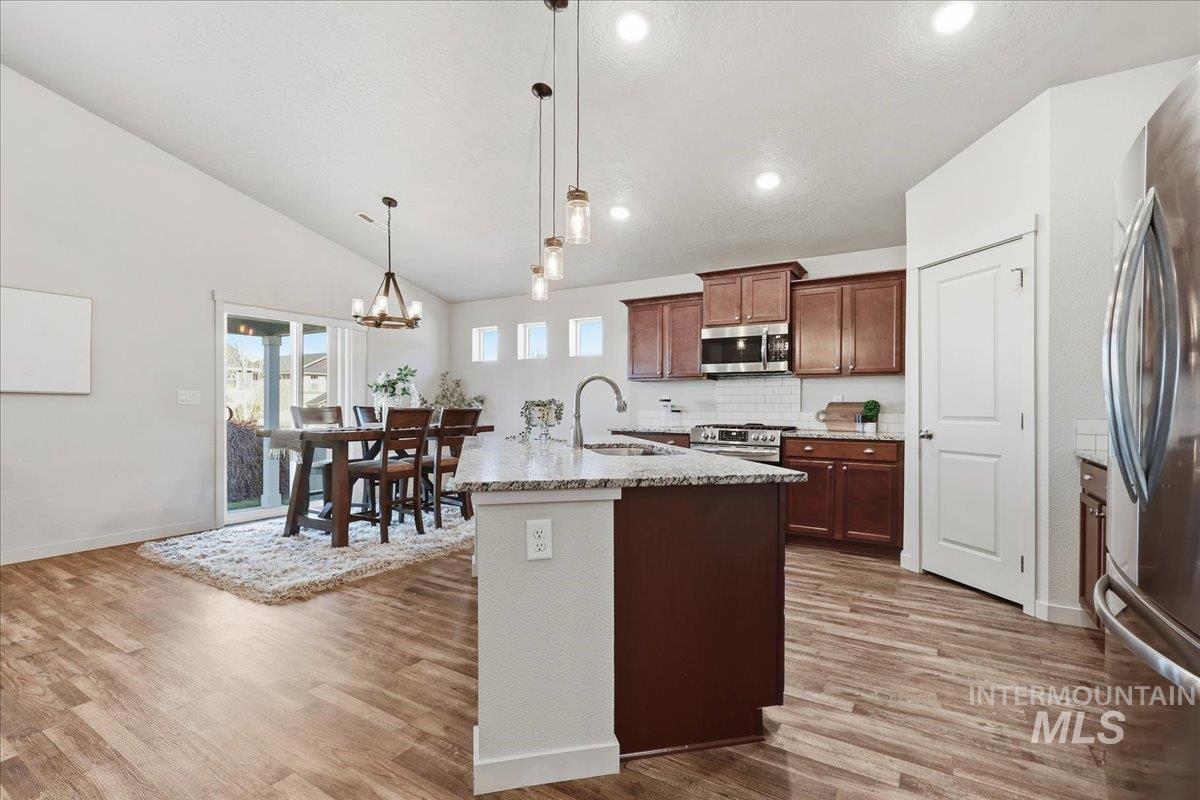 Kitchen featuring appliances with stainless steel finishes, a center island with sink, decorative light fixtures, light stone counters, and backsplash