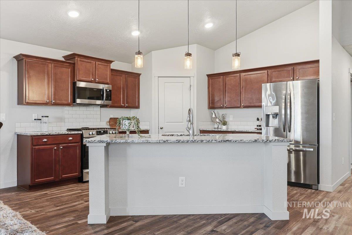 Kitchen featuring appliances with stainless steel finishes, vaulted ceiling, tasteful backsplash, light stone counters, and pendant lighting