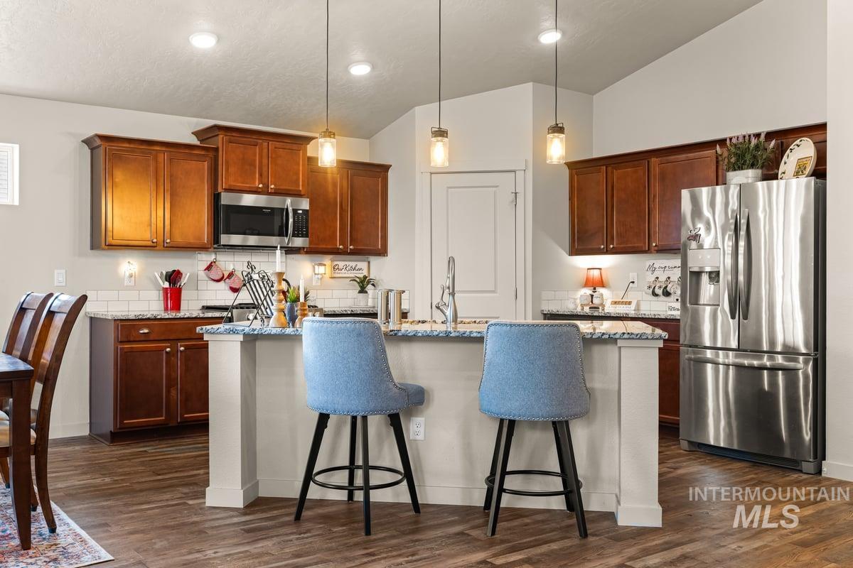 Kitchen featuring a kitchen island with sink, stainless steel appliances, light stone countertops, hanging light fixtures, and dark wood-style floors