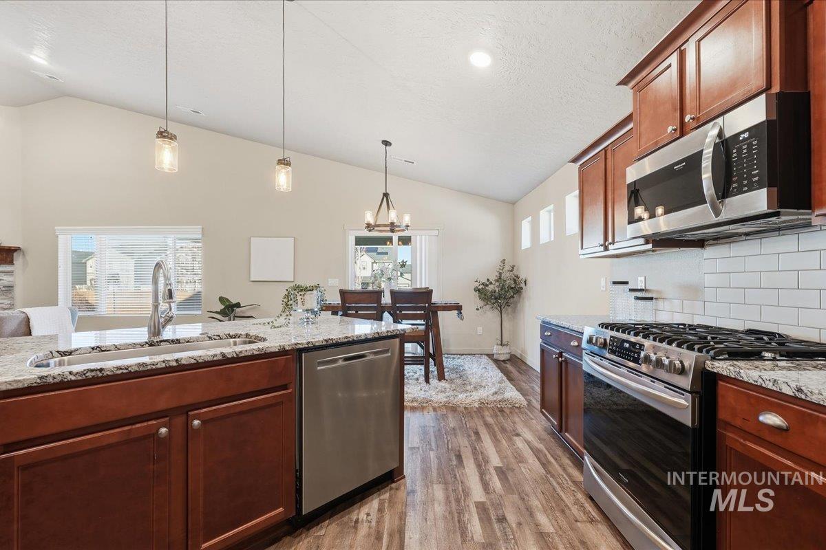 Kitchen featuring stainless steel appliances, lofted ceiling, decorative light fixtures, light wood-style floors, and light stone countertops