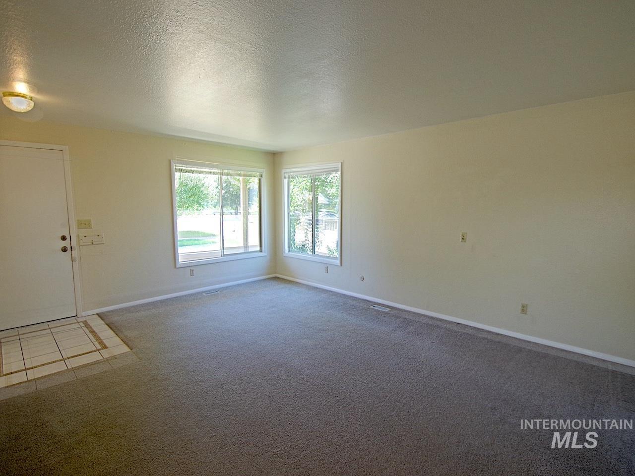 Spacious room featuring light neutral wall paint, two large windows, grey carpeting, a tiled entryway, and a white entry door