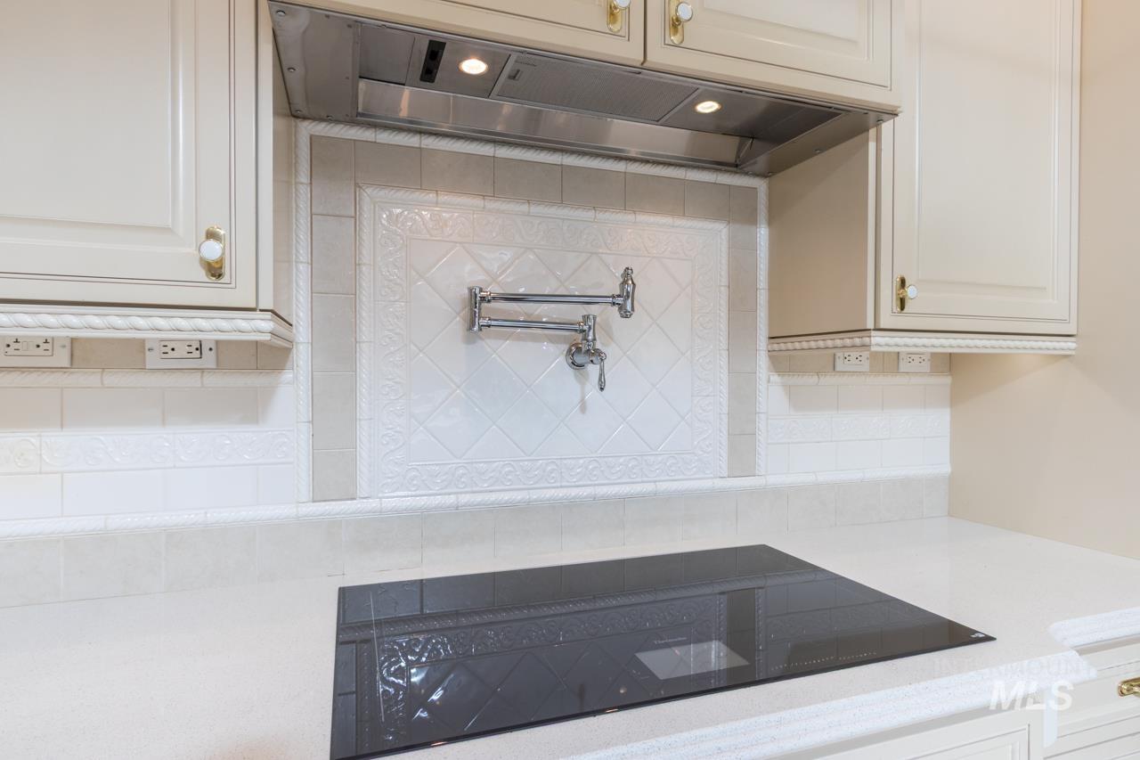 Kitchen view of black electric cooktop, pot filler, light stone countertops, backsplash, and white cabinetry
