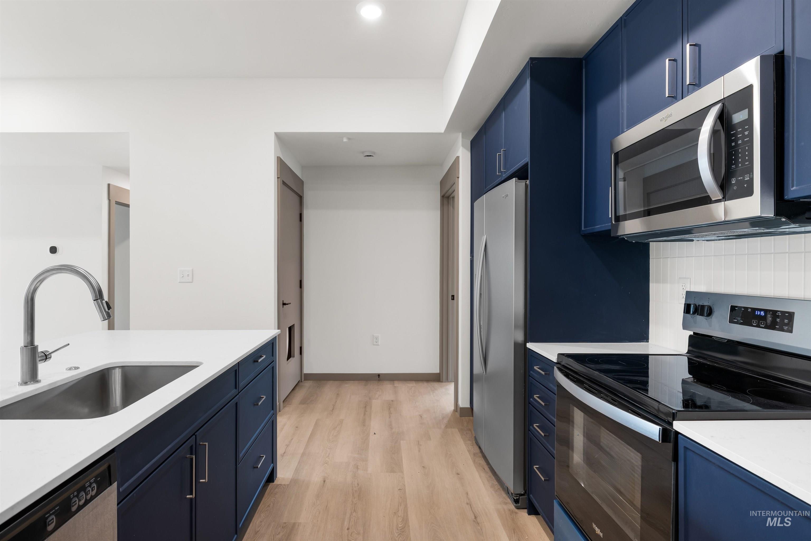 Kitchen featuring blue cabinetry, stainless steel appliances, light wood-type flooring, light stone countertops, and tasteful backsplash