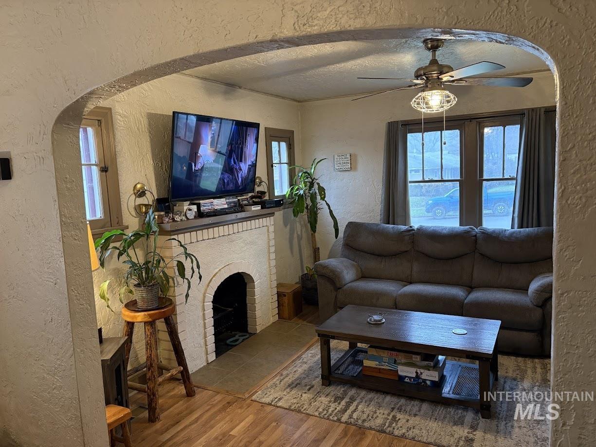 Living room featuring a textured wall, arched walkways, ceiling fan, wood finished floors, and a brick fireplace