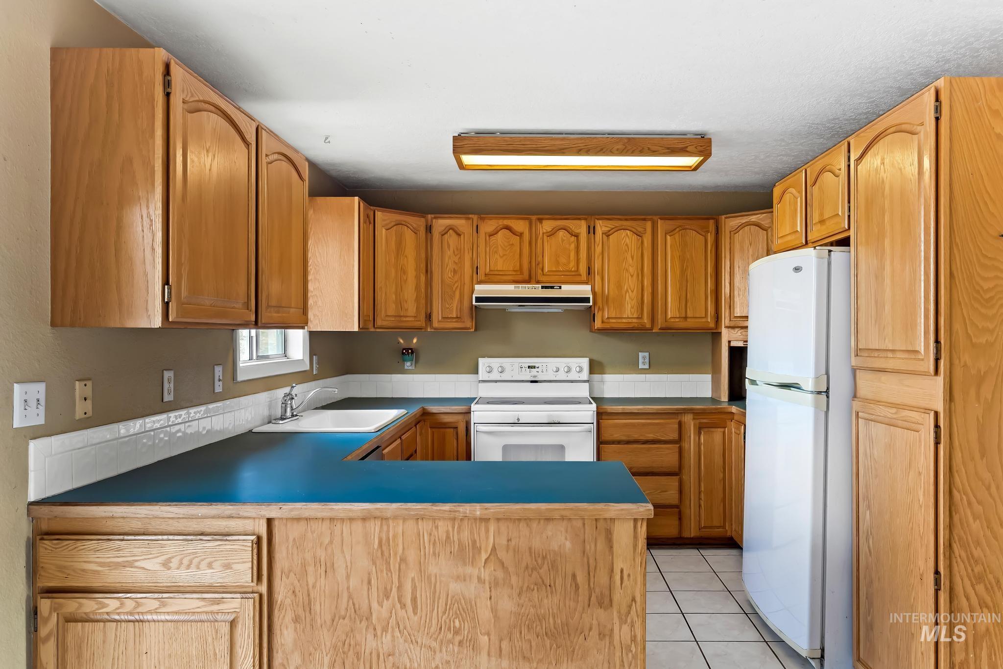 Kitchen with white appliances, light tile patterned floors, a peninsula, brown cabinets, and under cabinet range hood