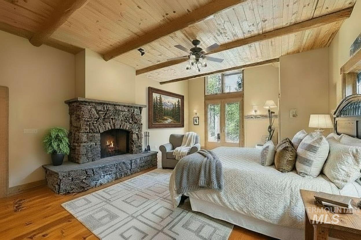 Bedroom with a wooden ceiling with exposed beams, wood finished floors, a stone fireplace, and ceiling fan
