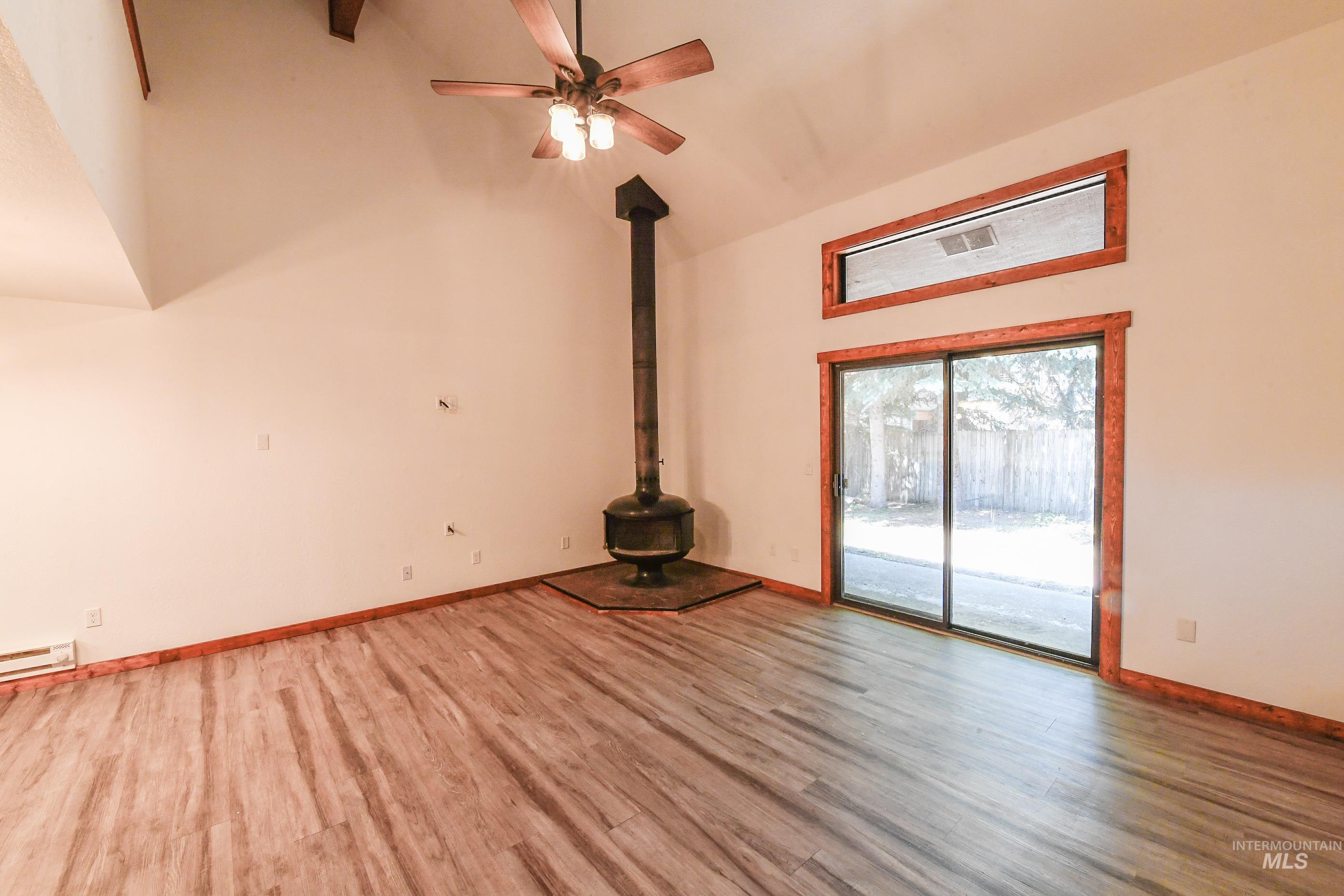 Unfurnished living room featuring a wood stove, wood finished floors, high vaulted ceiling, ceiling fan, and a baseboard radiator