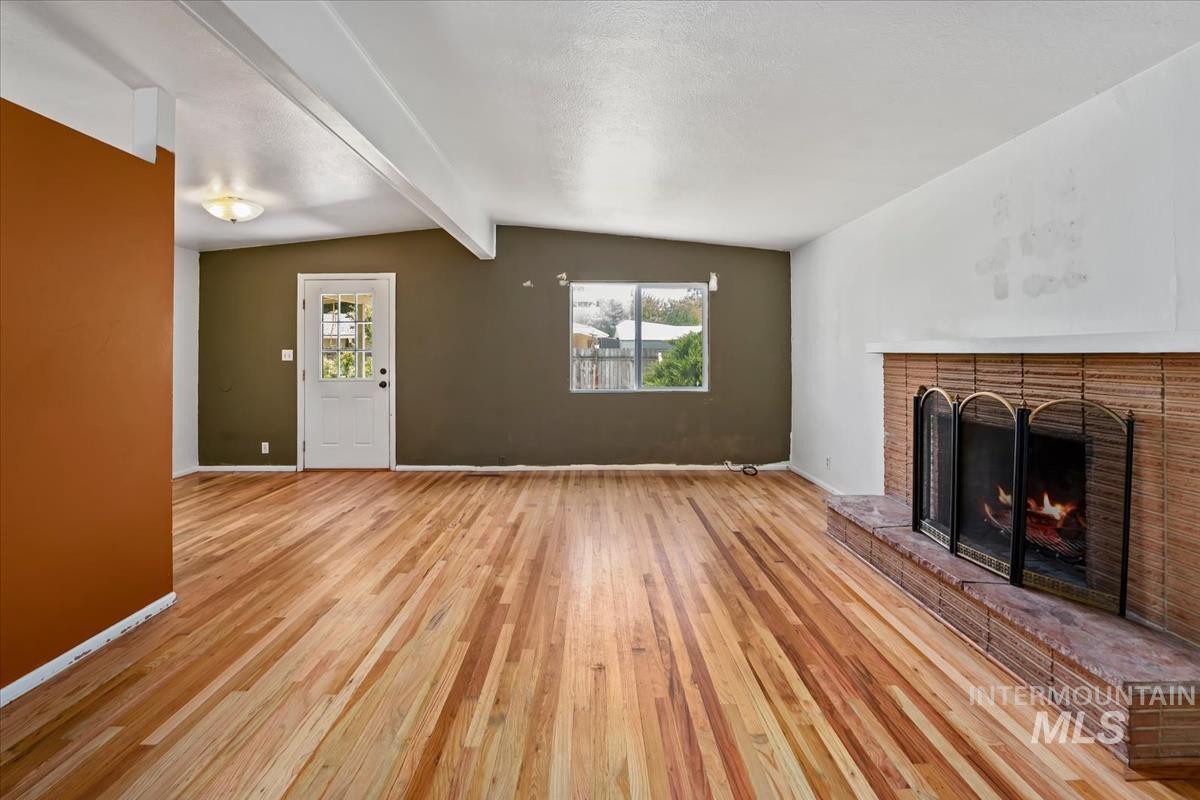 Living area featuring wood-finish flooring, a brick fireplace with a hearth, a vaulted ceiling with exposed beams, and a paneled entry door with glass inserts