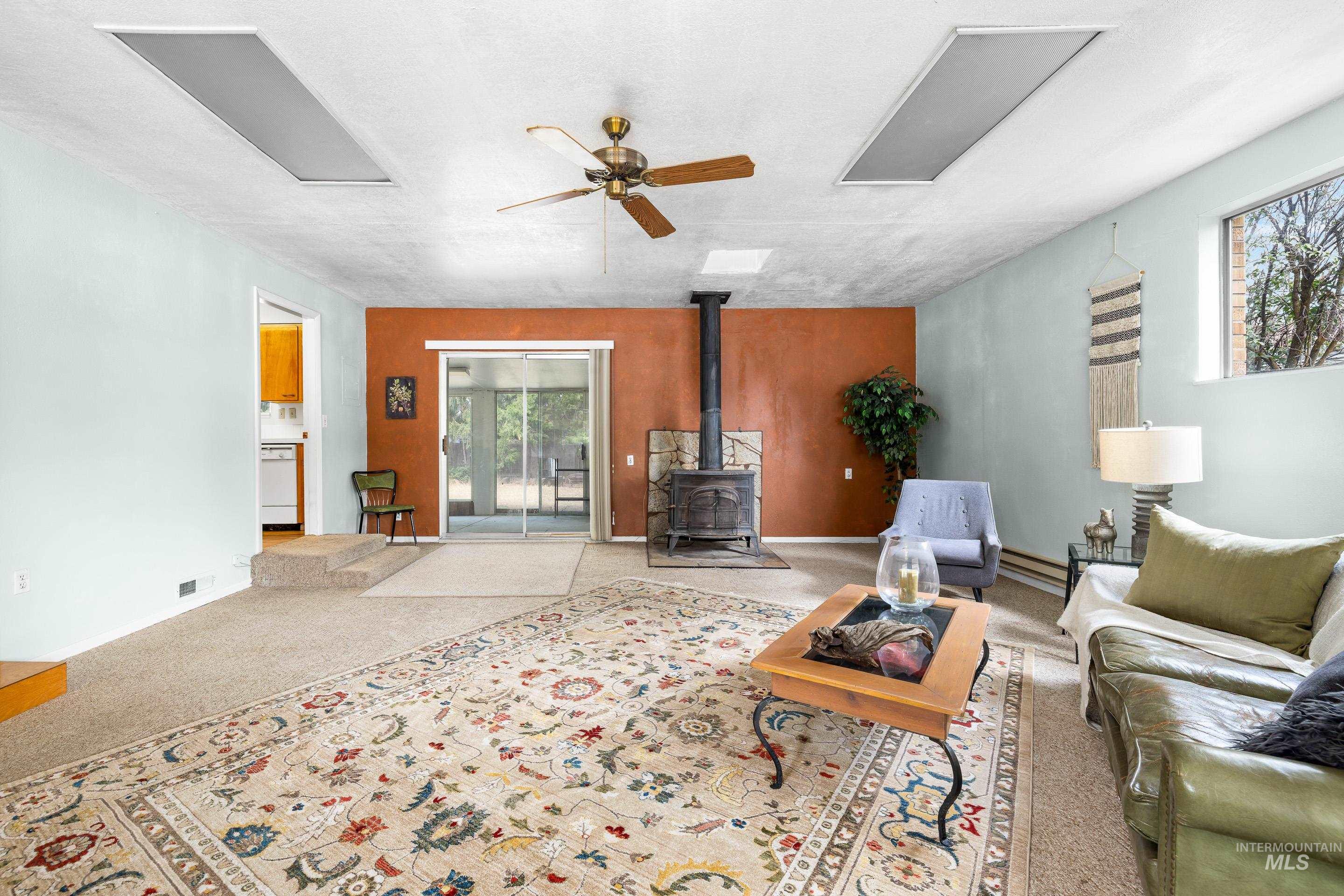 Carpeted living room featuring a wood stove, ceiling fan, a baseboard heating unit, and a textured ceiling