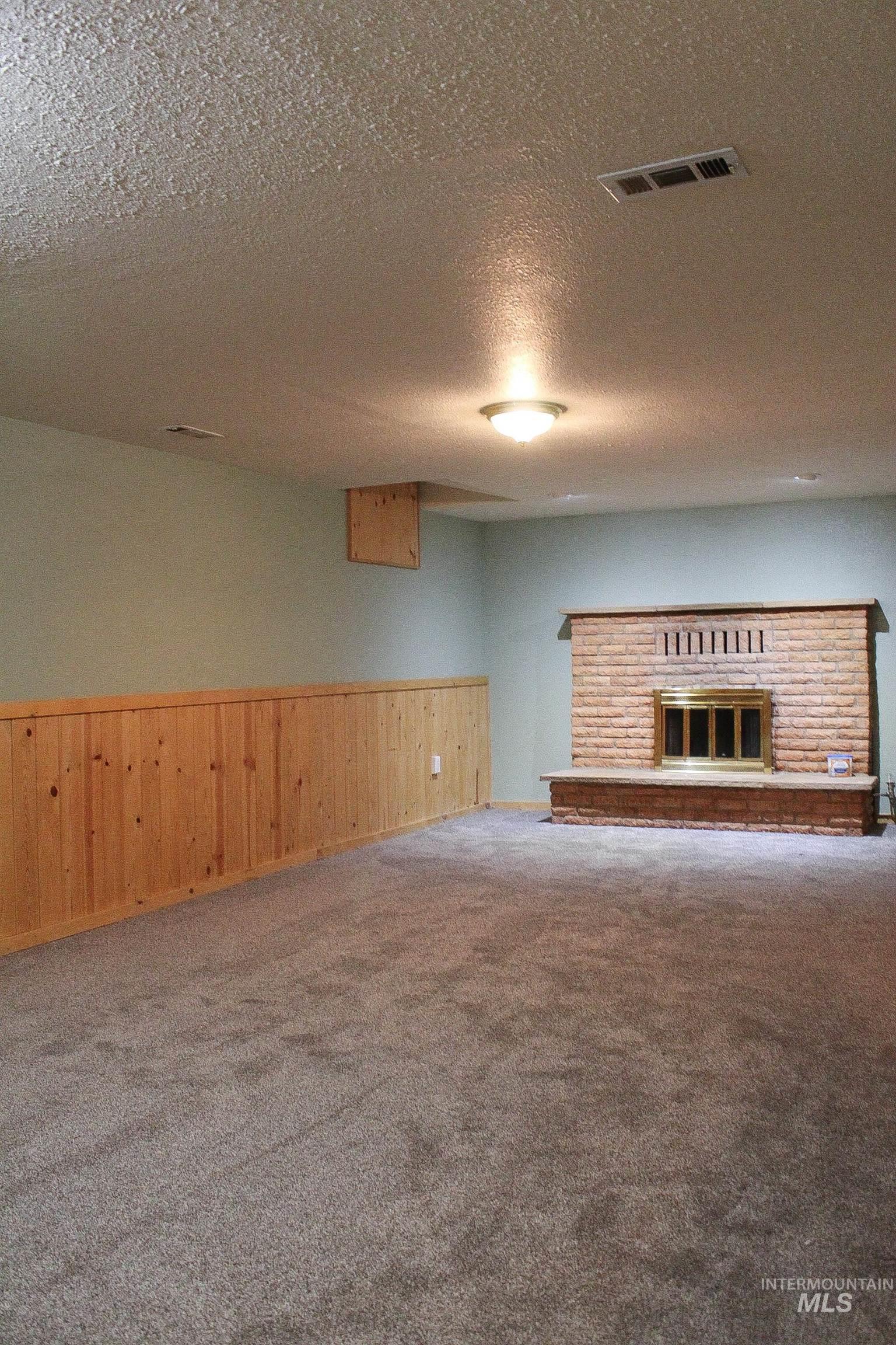 Unfurnished living room with carpet flooring, a textured ceiling, a brick fireplace, a wainscoted wall, and wood walls