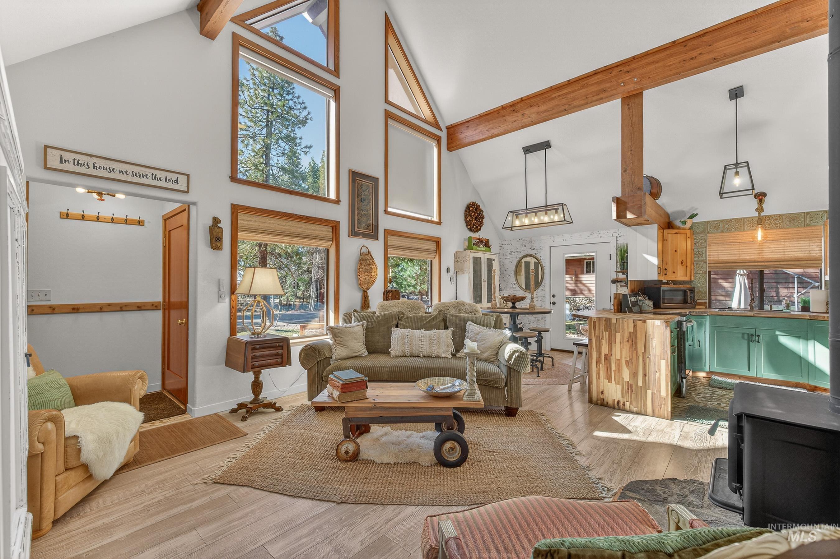 Vaulted ceiling with exposed wood beams, wood-finish flooring, multiple oversized windows, built-in shelving, and a prominent wood-burning stove
