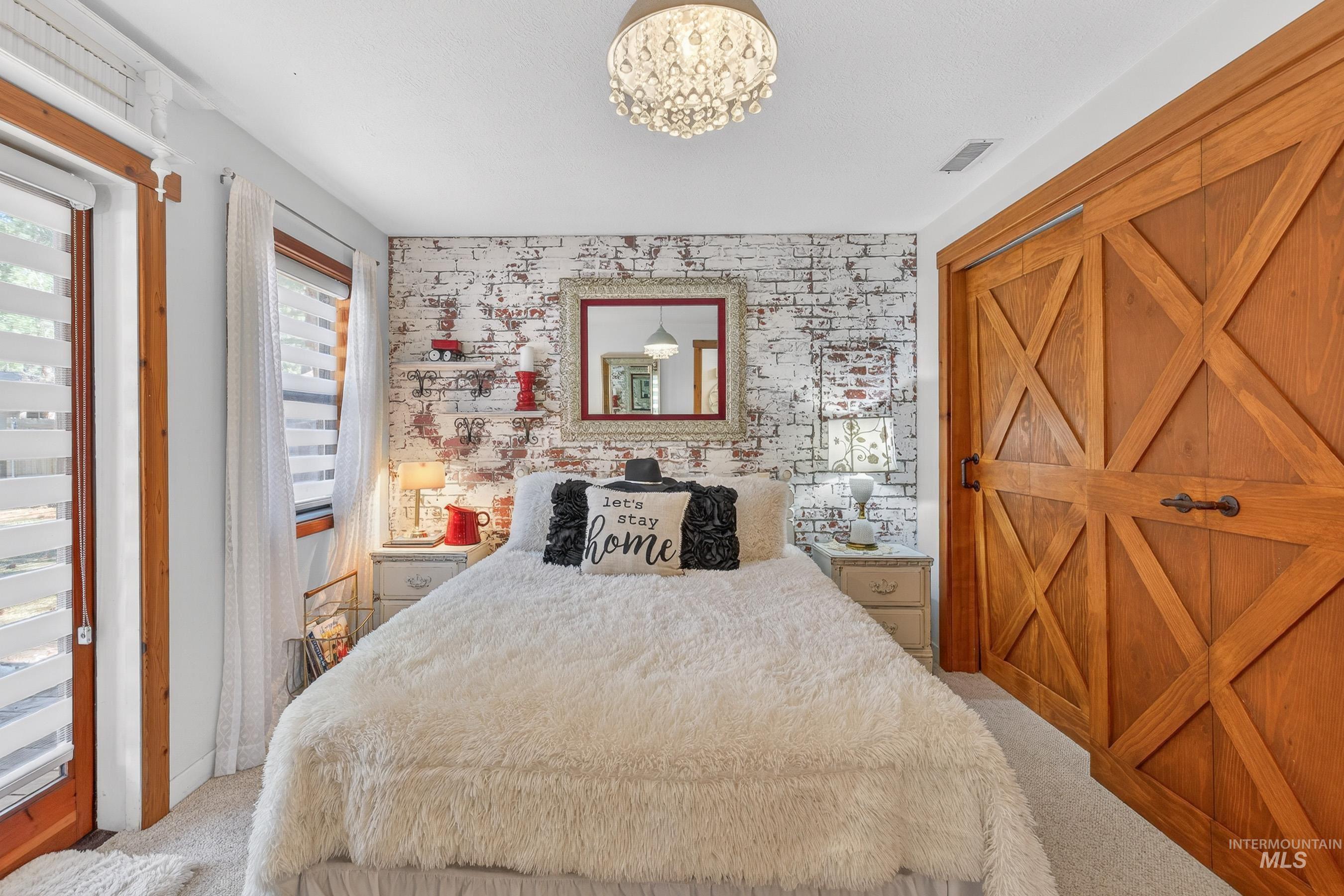 Bedroom featuring a decorative chandelier, white-washed brick accent wall, wood-frame windows, and large wood sliding barn doors
